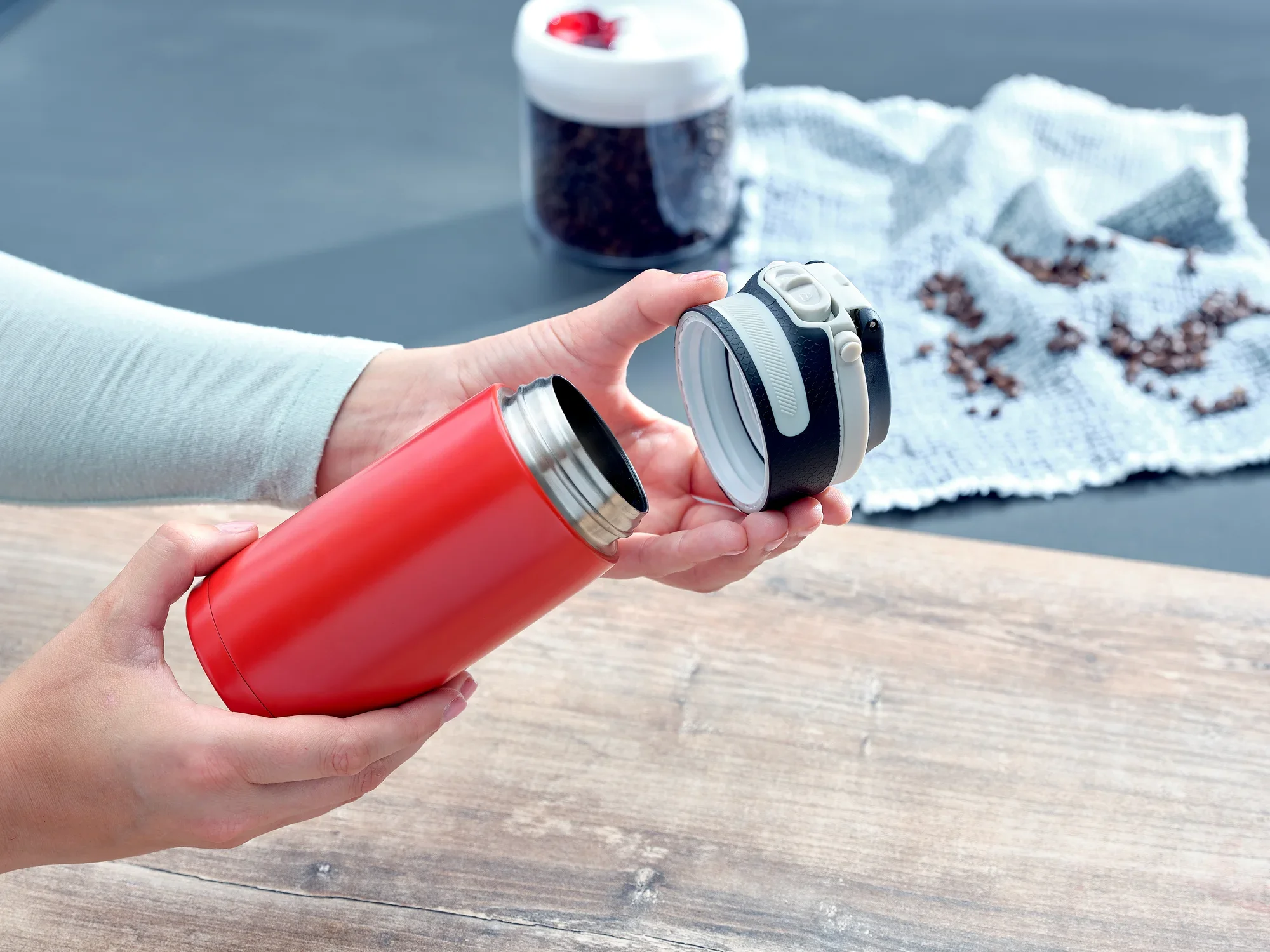 Czerwony izolowany kubek Flip 350ml otwierany przez osobę. W tle widać ziarna kawy i białą ściereczkę. Idealny do utrzymywania ciepła napojów podczas podróży.