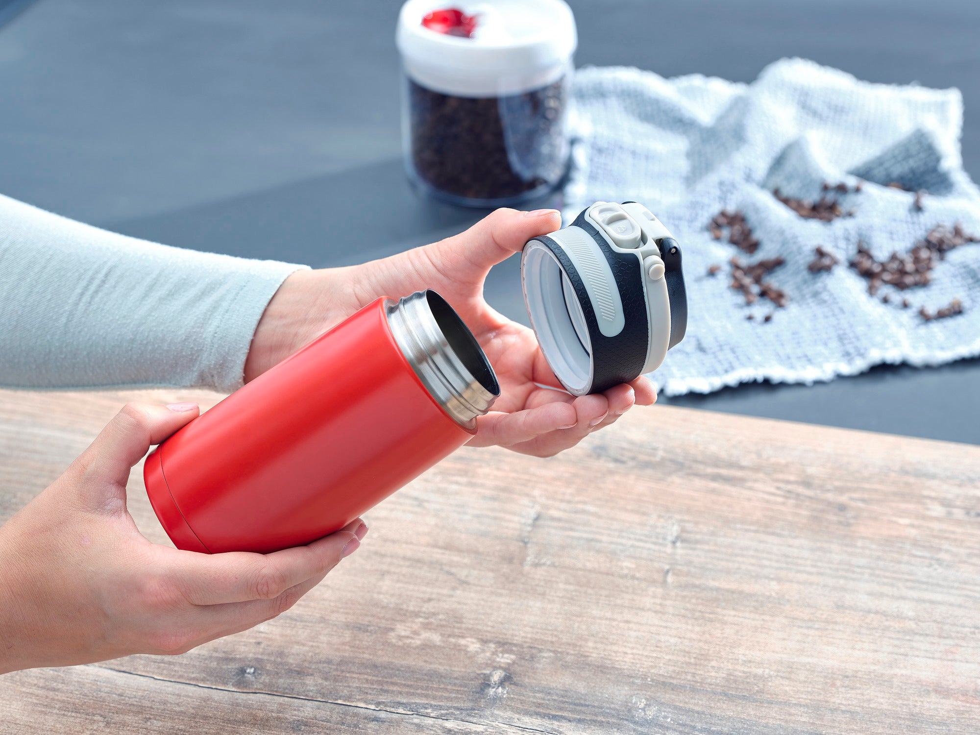 Czerwony izolowany kubek Flip 350ml otwierany przez osobę. W tle widać ziarna kawy i białą ściereczkę. Idealny do utrzymywania ciepła napojów podczas podróży.