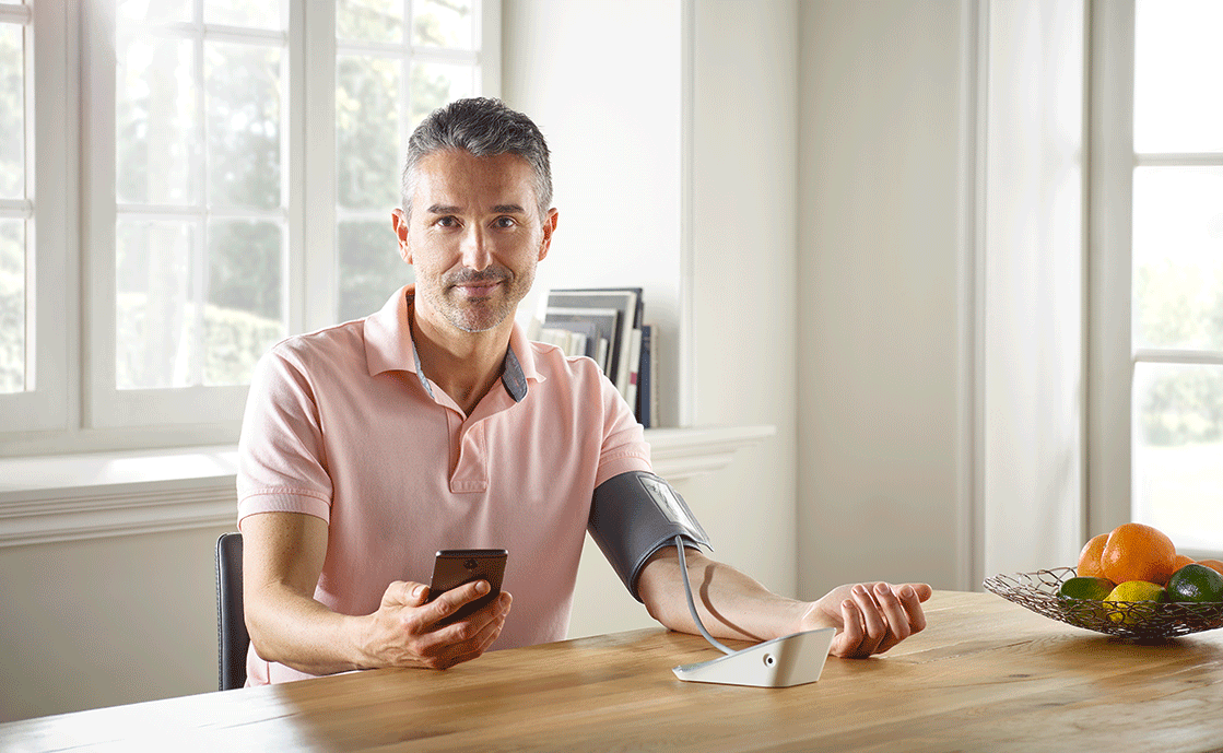 Ein Mann in rosa Polohemd misst seinen Blutdruck mit Smartphone und Blutdruckmessgeraet an einem Tisch mit Obstschale.
