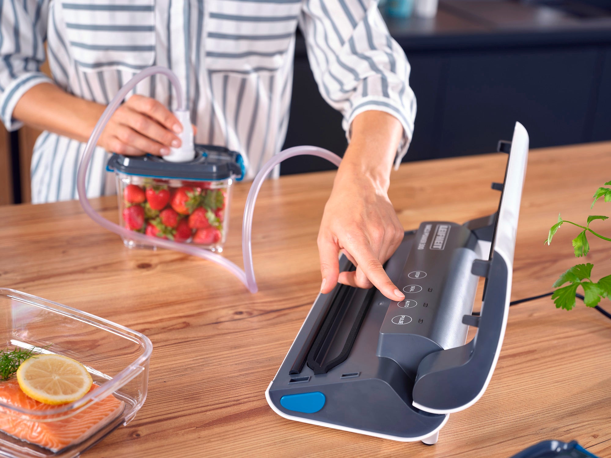 Une personne utilise l'appareil de mise sous vide Vacu Power 300 pour sceller des fraises fraîches dans un sachet. L'appareil est posé sur une table en bois à côté d'un plat contenant du saumon.