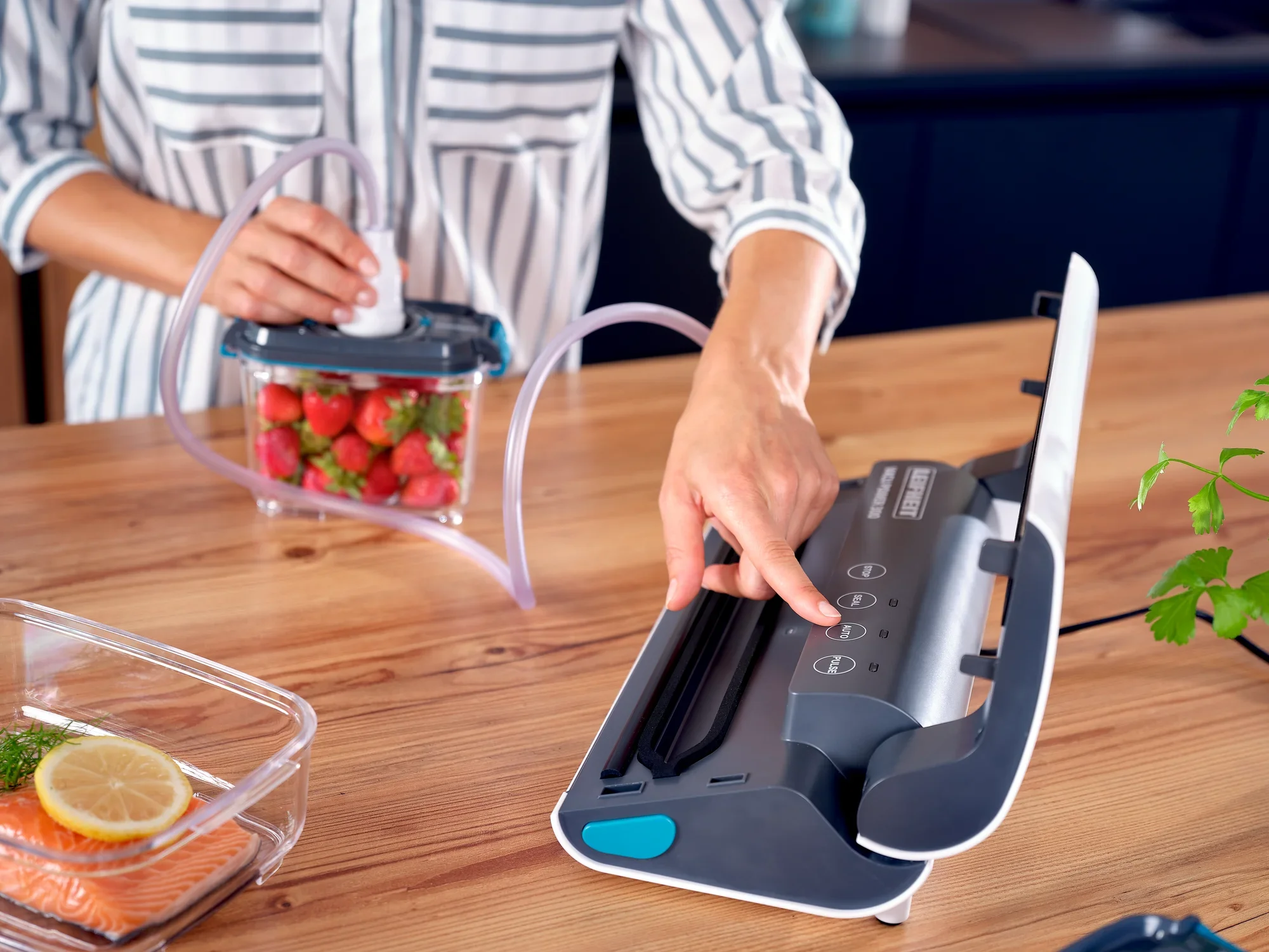 Une personne utilise l'appareil de mise sous vide Vacu Power 300 pour sceller des fraises fraîches dans un sachet. L'appareil est posé sur une table en bois à côté d'un plat contenant du saumon.