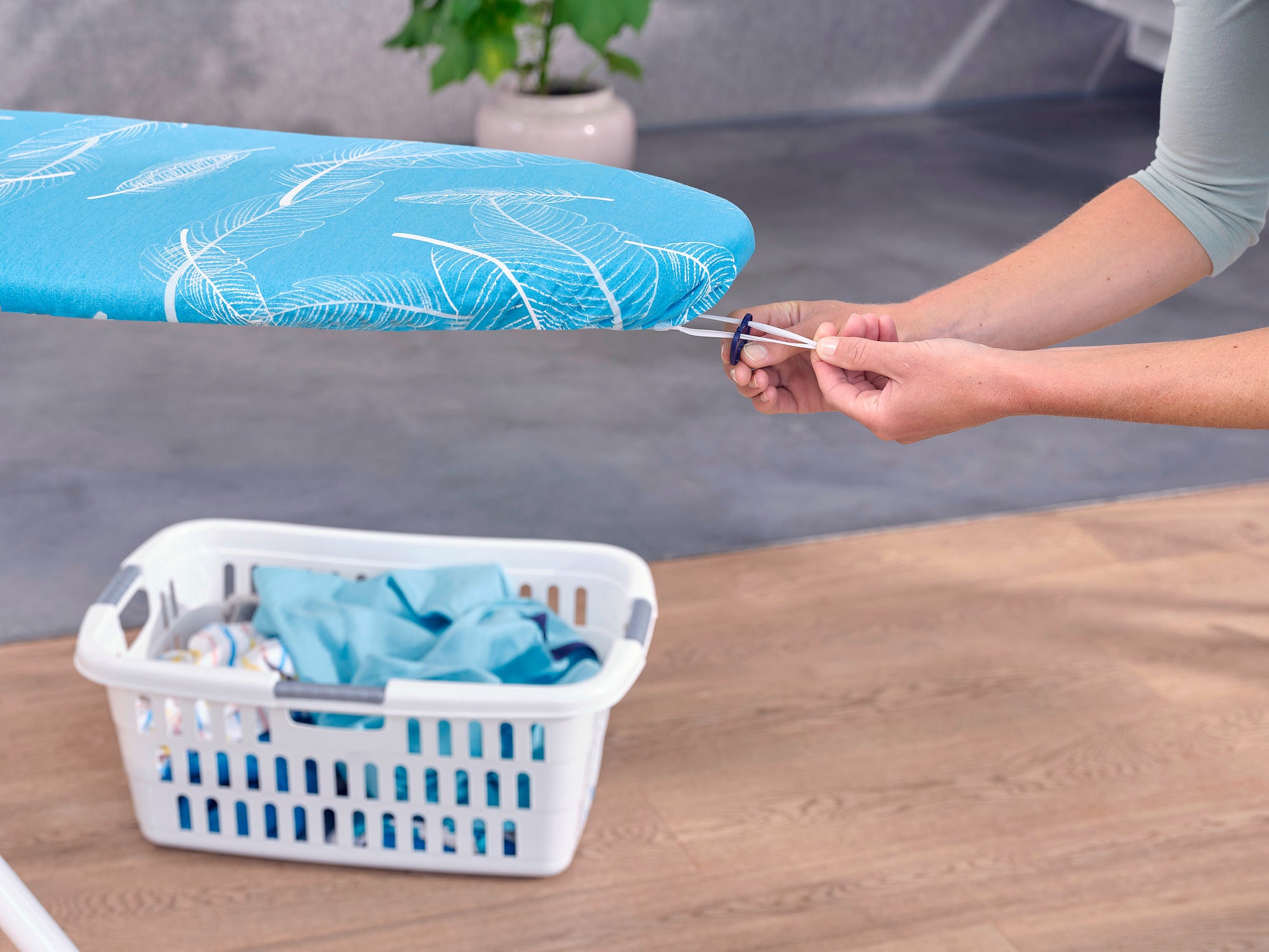 Strijkplank met een Thermo Reflect strijkplankovertrek in maat M. Een persoon trekt de hoes strak. Op de achtergrond staat een wasmand op een houten vloer.