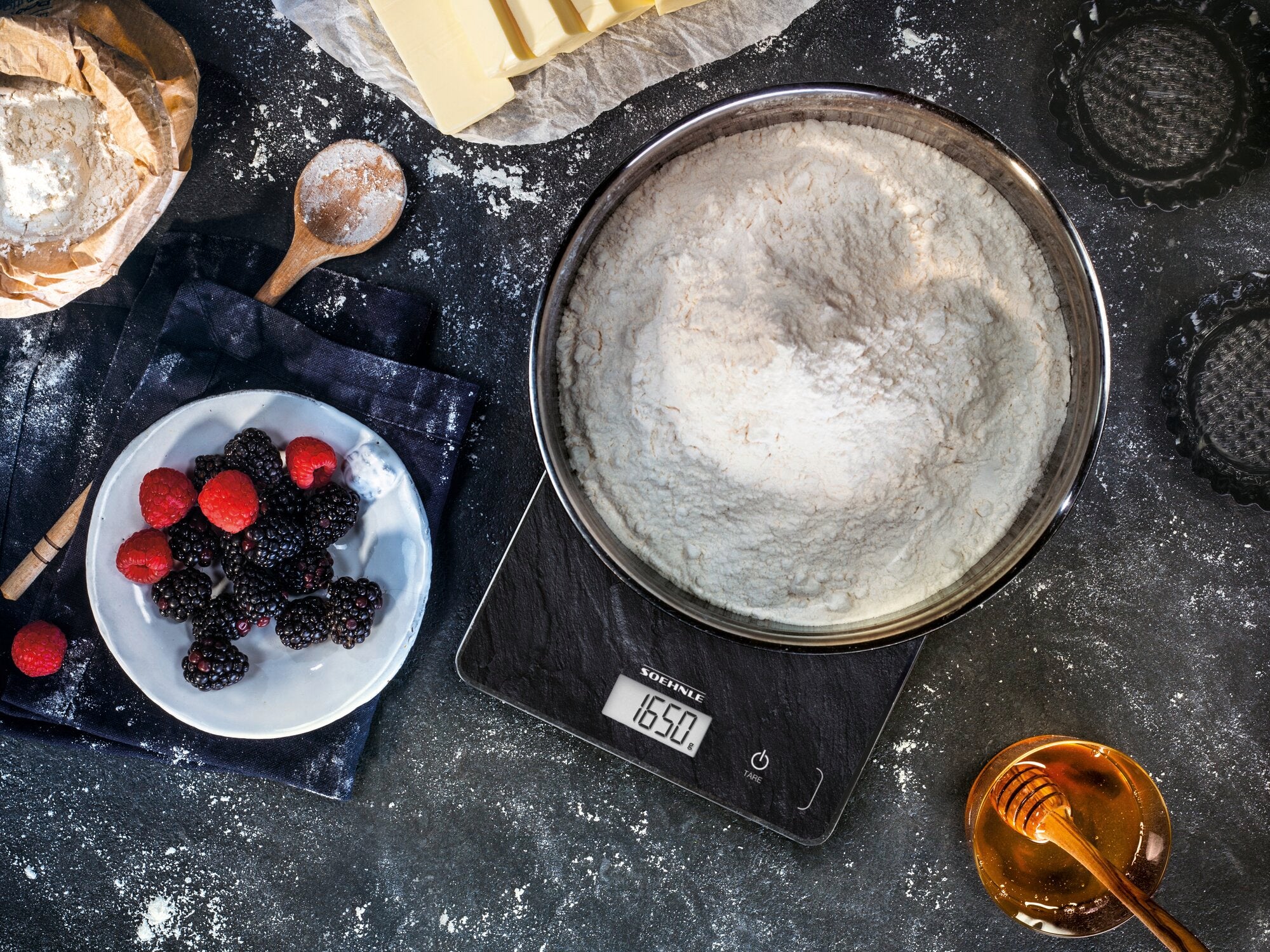 Cyfrowa waga kuchenna Page Compact 300 w kolorze łupkowym z miską mąki na powierzchni ważenia. Obok znajdują się jagody, miód i składniki do pieczenia.