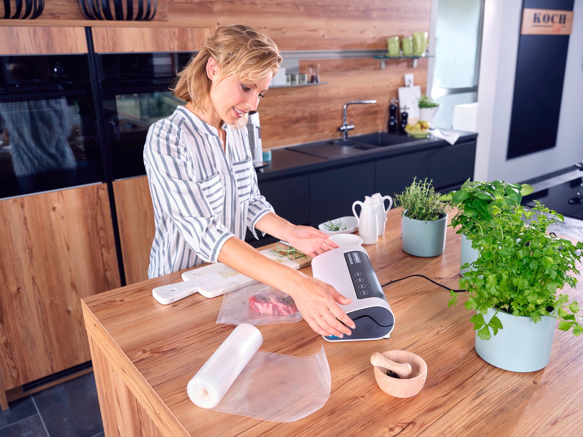 Une femme utilise l'appareil de mise sous vide Vacu Power 300 dans une cuisine moderne. Des herbes fraîches et des aliments sous vide sont posés sur le plan de travail. Idéal pour conserver la fraîcheur.