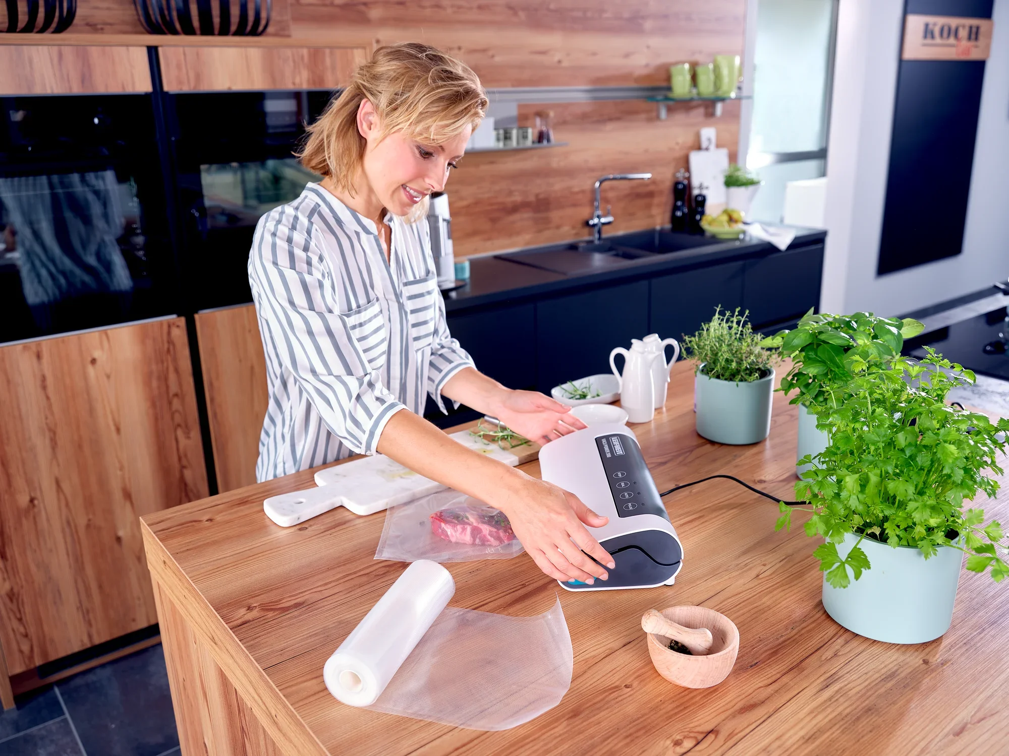 Une femme utilise l'appareil de mise sous vide Vacu Power 300 dans une cuisine moderne. Des herbes fraîches et des aliments sous vide sont posés sur le plan de travail. Idéal pour conserver la fraîcheur.