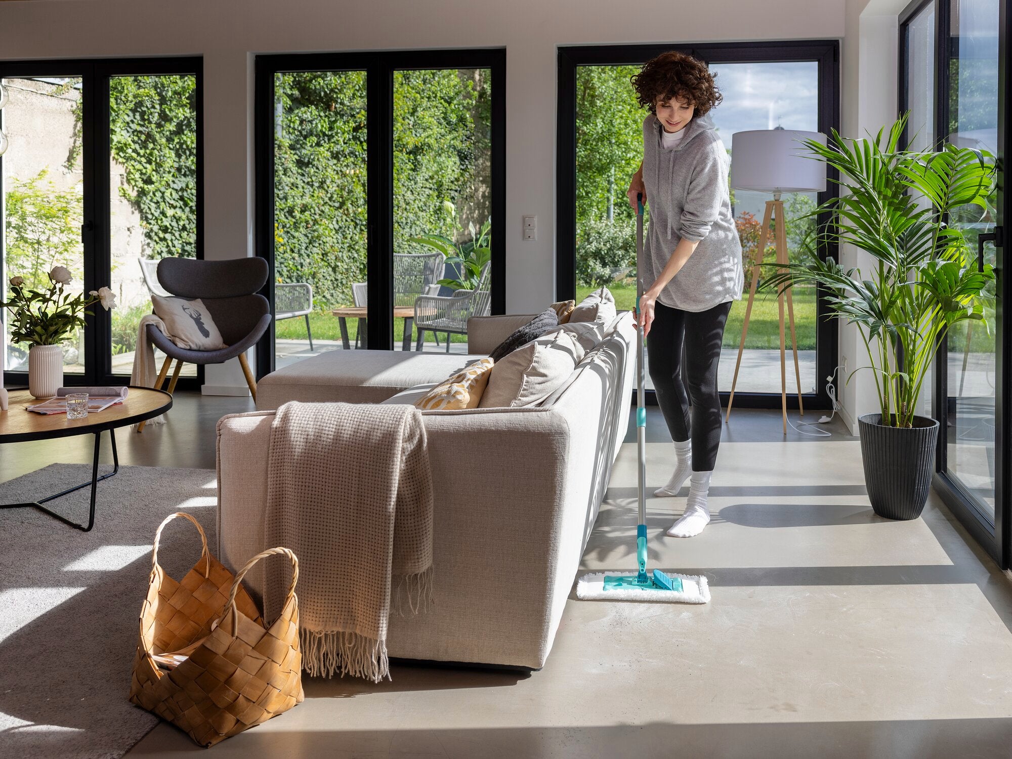Une personne nettoie le sol d'un salon moderne avec le plumeau SUPERDUSTER. De grandes fenêtres laissent entrer beaucoup de lumière, tandis que les plantes et les meubles créent une atmosphère chaleureuse.