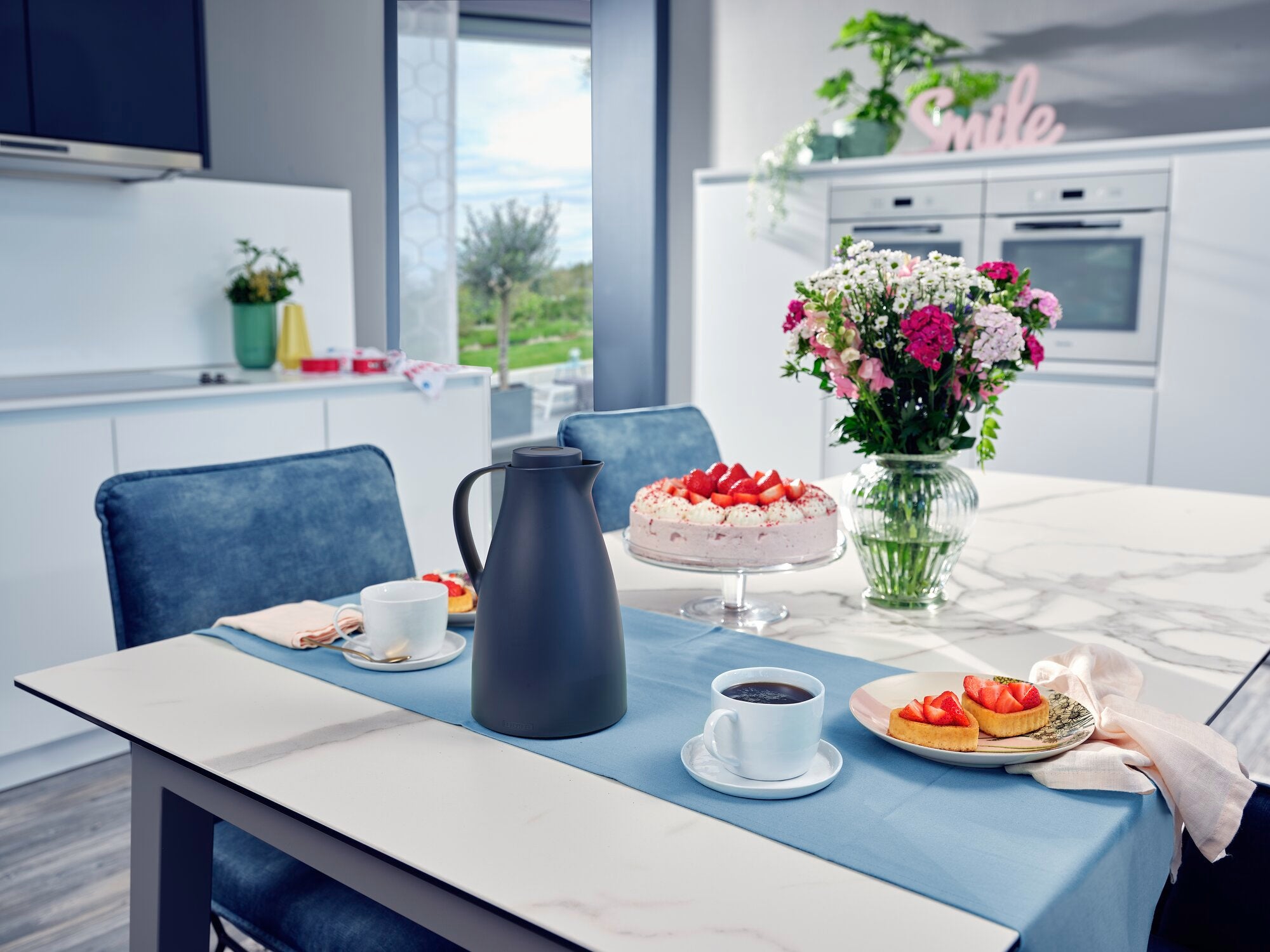 Eine stilvolle Küche mit einer Isolierkanne Harmonic auf einem Tisch. Daneben stehen Tassen mit Kaffee, ein Teller mit Erdbeerkuchen und ein Blumenstrauß. Im Hintergrund sind moderne Küchengeräte sichtbar.