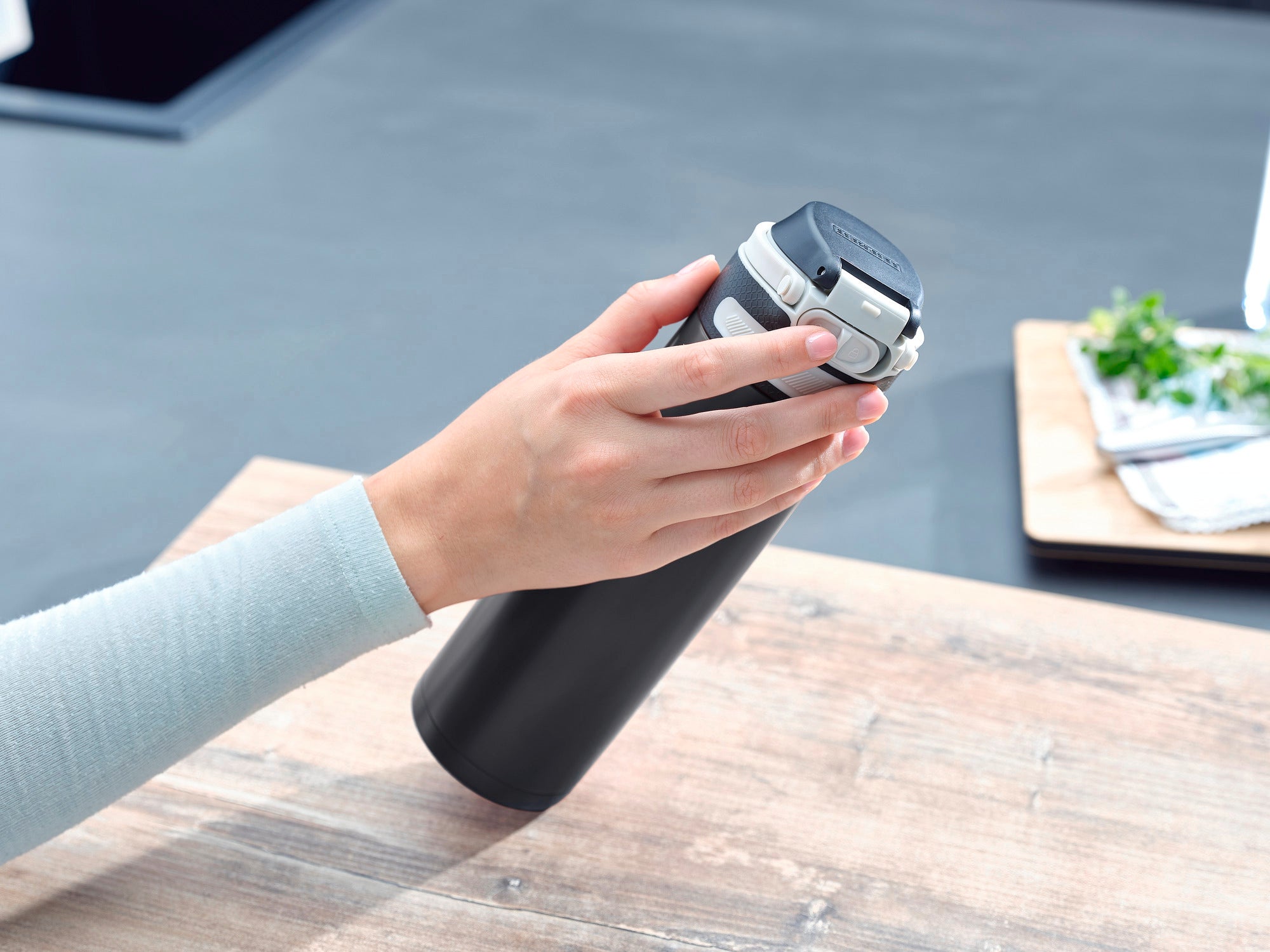 Une main tient un mug isotherme Flip noir d'une contenance de 600 ml. Le mug est posé sur une table en bois dans une cuisine moderne. Des herbes sont visibles à l'arrière-plan.
