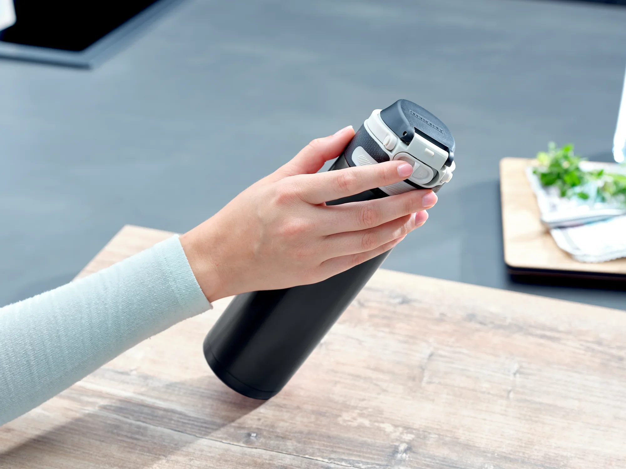 Jedna ręka trzyma czarny kubek próżniowy Flip o pojemności 600 ml. Kubek stoi na drewnianym stole w nowoczesnej kuchni. W tle widoczne są zioła.