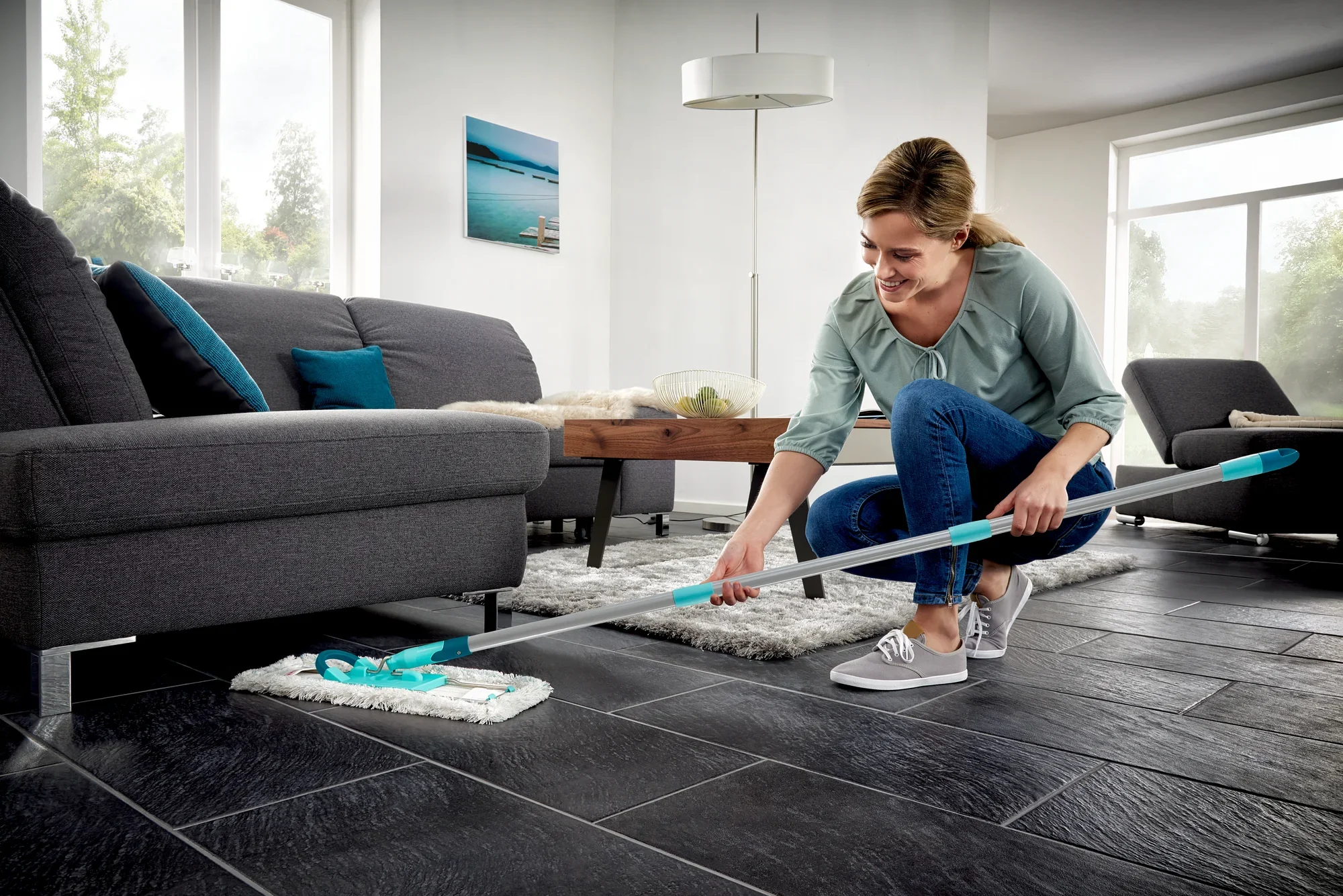 Eine Frau reinigt den Boden eines modernen Wohnzimmers mit dem Bodenwischer Profi XL. Der Wischer hat einen langen Stiel und ein Baumwollpad, ideal für große Flächen.