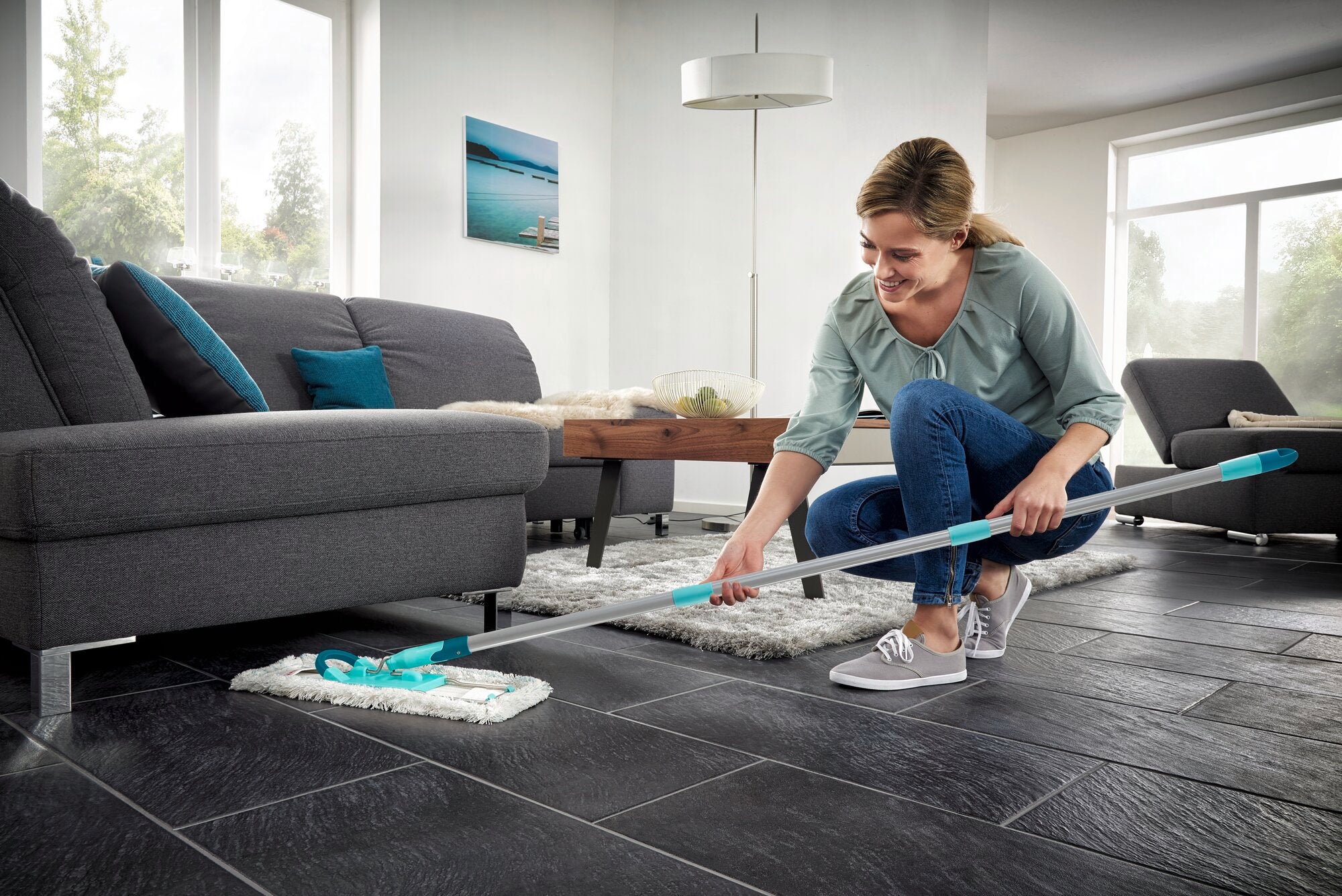 Kobieta myje podłogę w nowoczesnym salonie za pomocą ściągaczki do podłóg Profi XL. Mop ma długi uchwyt i bawełnianą nakładkę, idealną do dużych powierzchni.