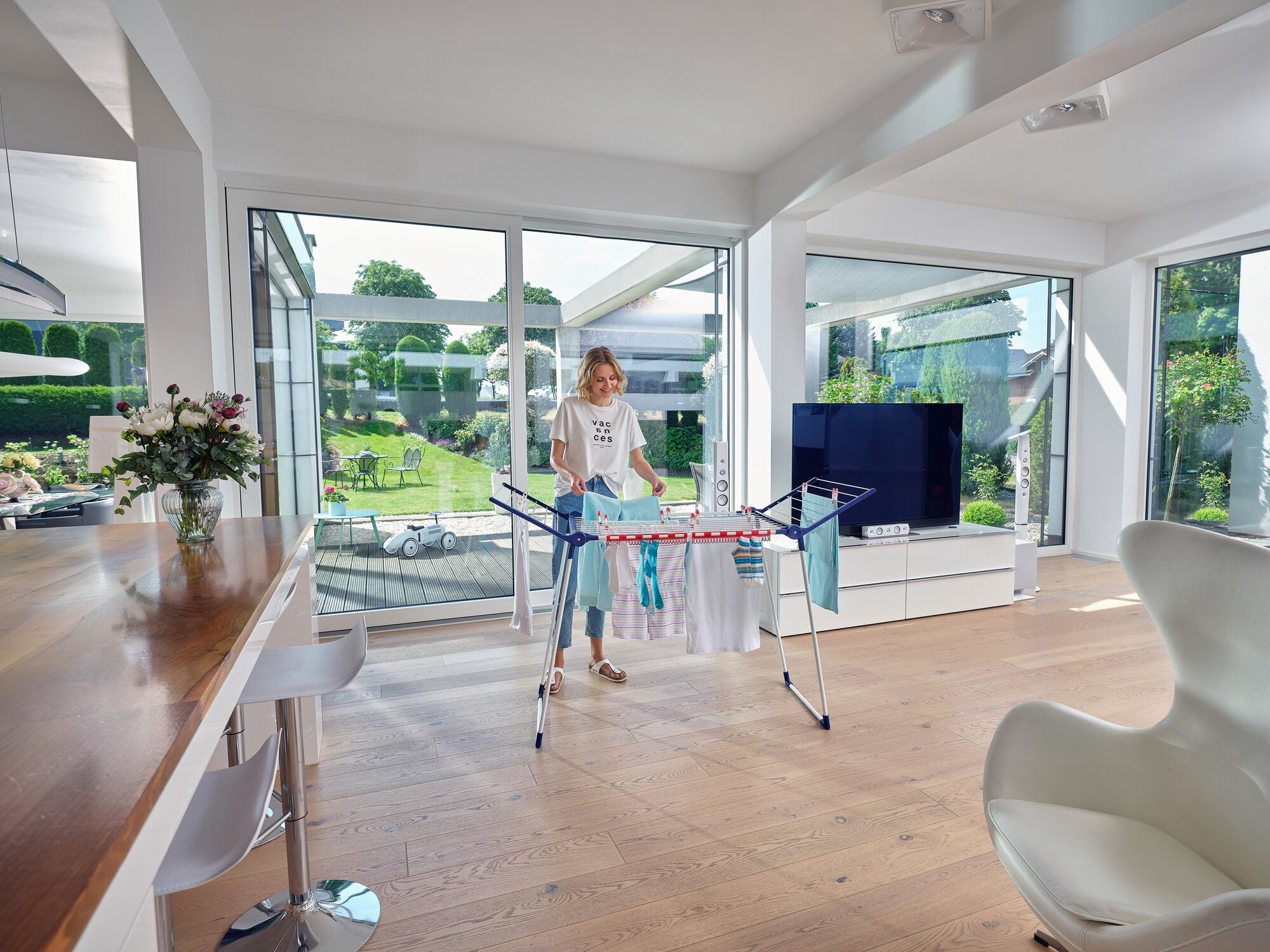 A woman stands in a modern, bright living room using the Pegasus 180 Solid Classic free-standing dryer. The dryer is covered with colourful laundry.