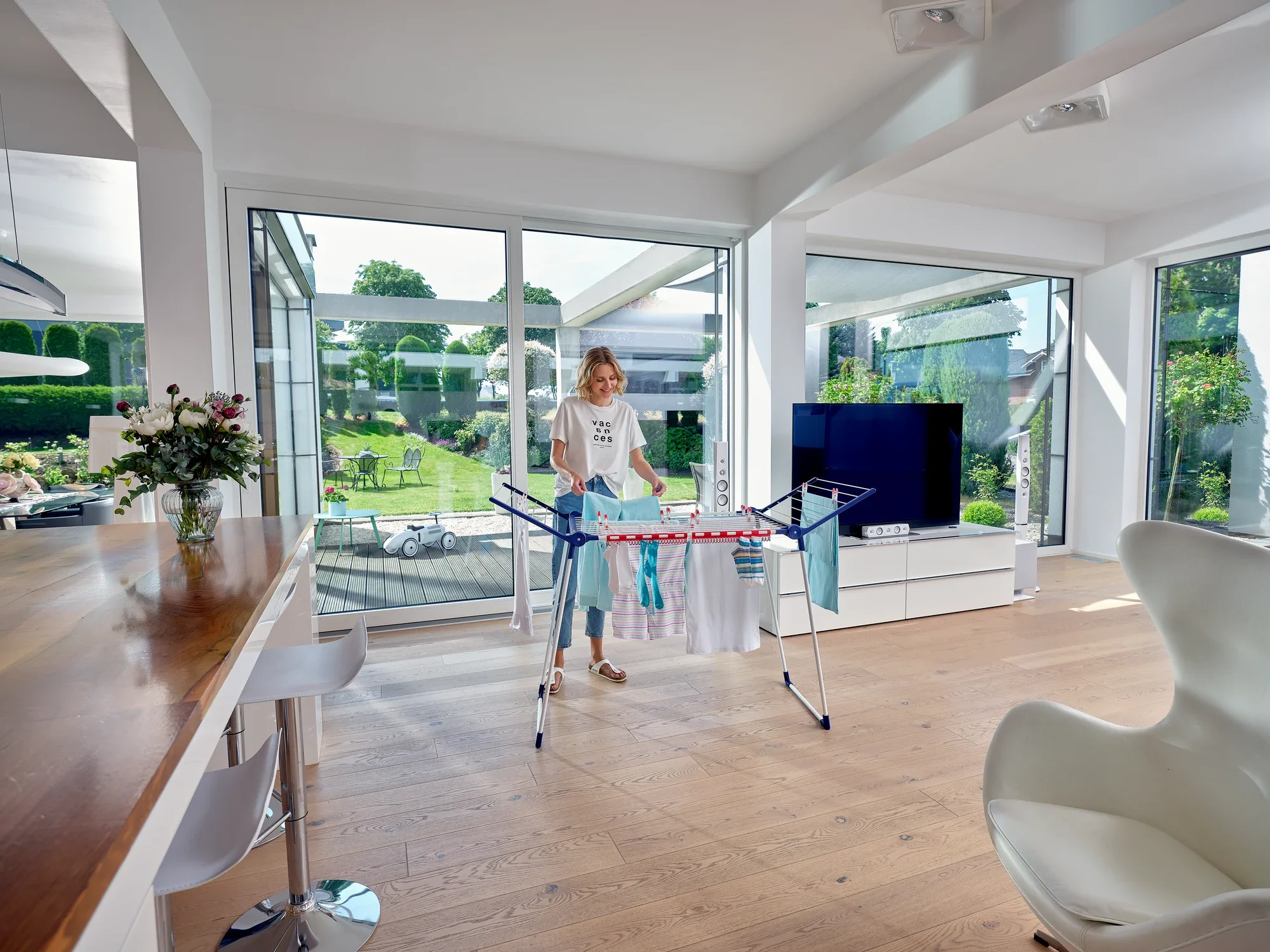 Kobieta stoi w nowoczesnym, jasnym salonie i korzysta z suszarki bębnowej Pegasus 180 Solid Classic. Na suszarce rozwieszone jest kolorowe pranie.