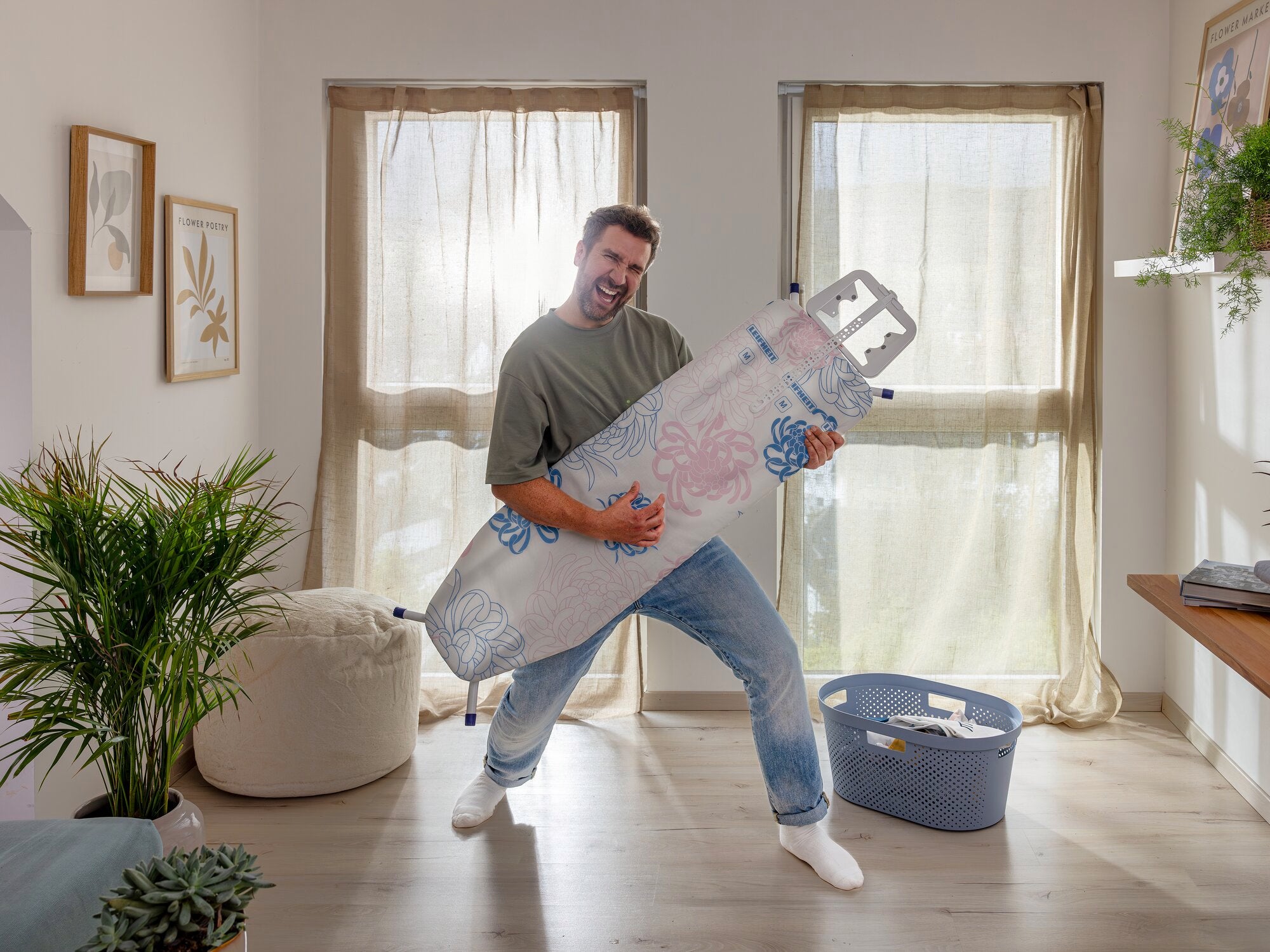 Ein Mann steht in einem hellen Raum und hält einen Bügeltisch Classic M Basic Plus wie eine Gitarre. Der Raum ist gemütlich eingerichtet mit Pflanzen und einem Wäschekorb.