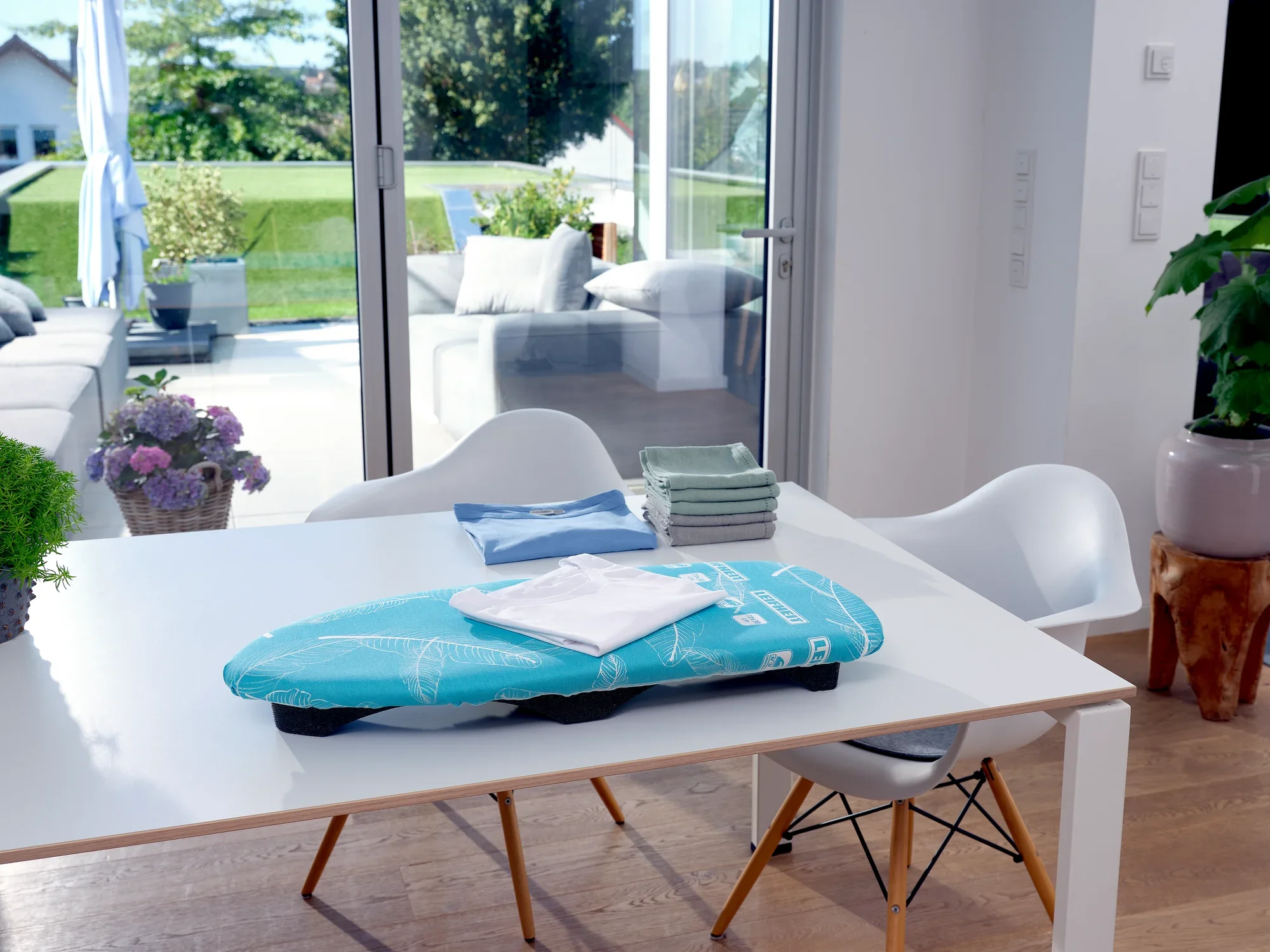 A compact ironing table, the Air Board Table Compact, lies on a modern dining table in a bright room with a view of a sunny terrace.
