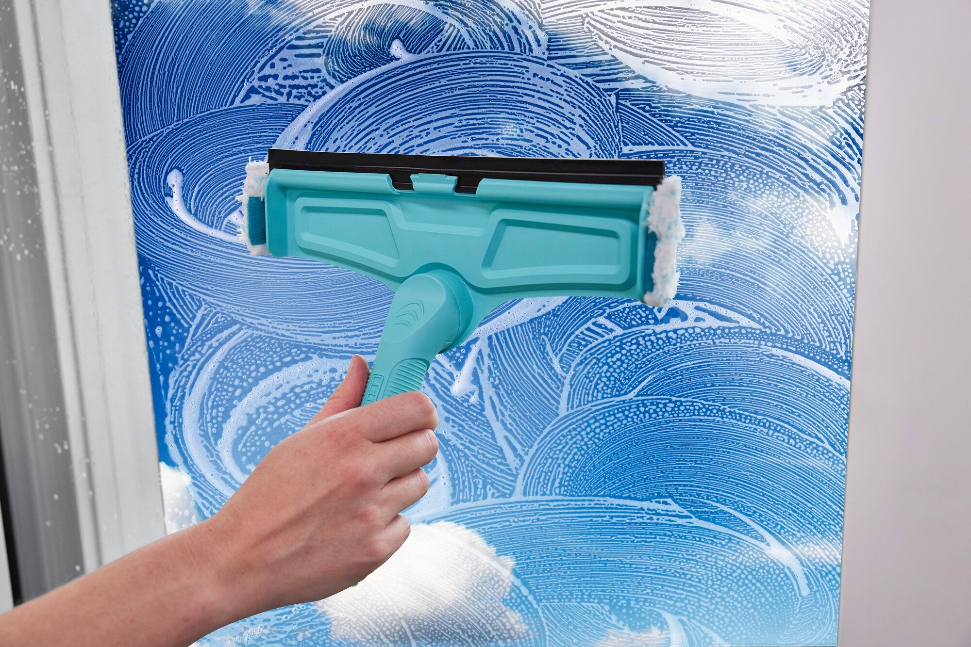 Un pulitore per finestre viene utilizzato per pulire una finestra con un detergente blu. La mano tiene il Window & Frame Cleaner 4in1, che rimuove efficacemente lo sporco.