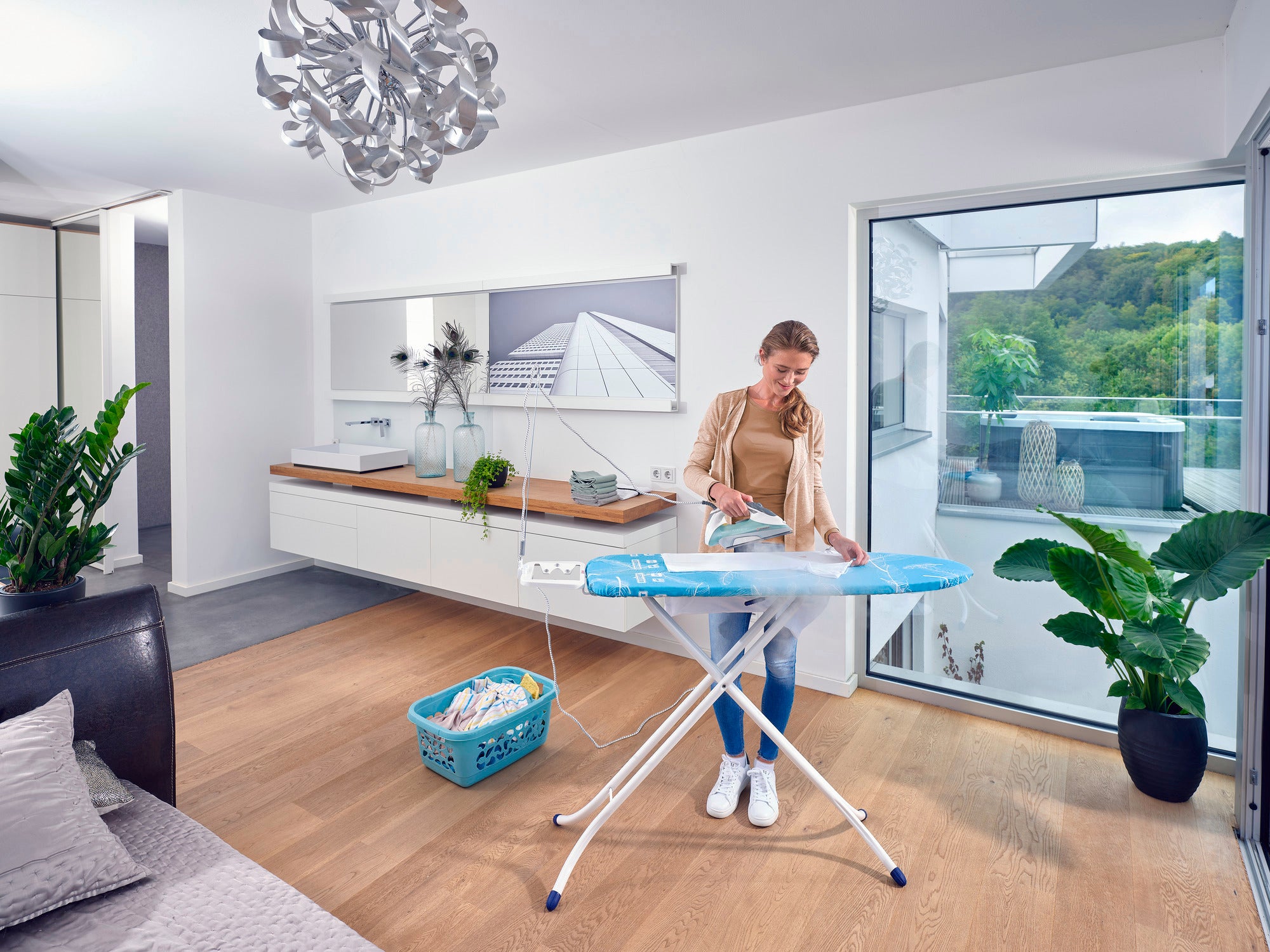 Eine Frau bügelt Kleidung auf einem Leifheit Air Board M Compact Bügeltisch in einem modernen, hellen Raum mit großen Fenstern und Pflanzen. Ein Wäschekorb steht daneben.