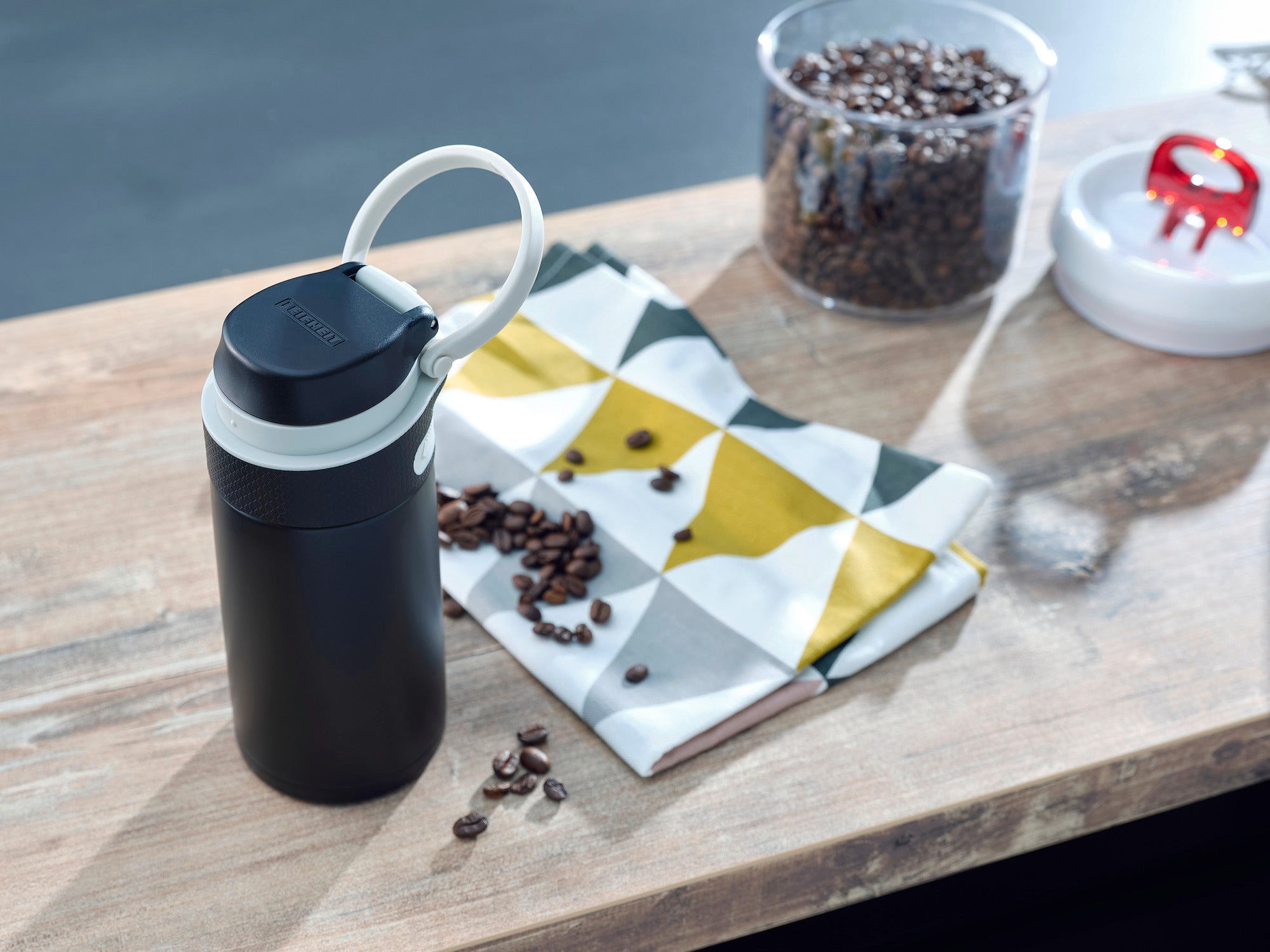 Un mug isotherme Flip noir d'une contenance de 350 ml est posé sur une table en bois. À côté se trouvent des grains de café et un tissu à motifs. À l'arrière-plan, on aperçoit un récipient contenant des grains de café.