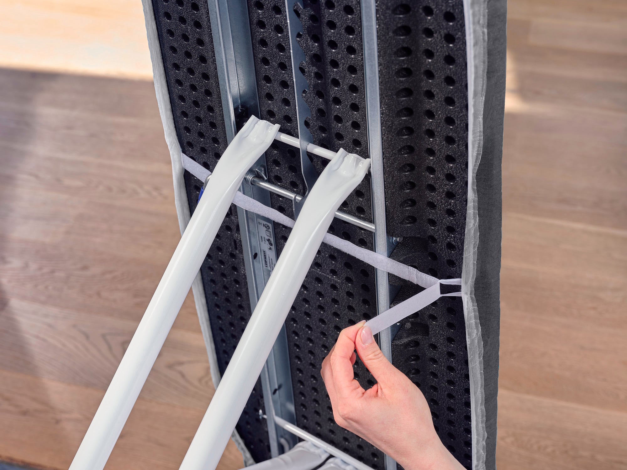 Tabla de planchar con la funda Thermo Reflect Black. Una mano tira de las bandas elásticas para fijar la funda de forma segura. La funda ofrece una superficie reflectante.