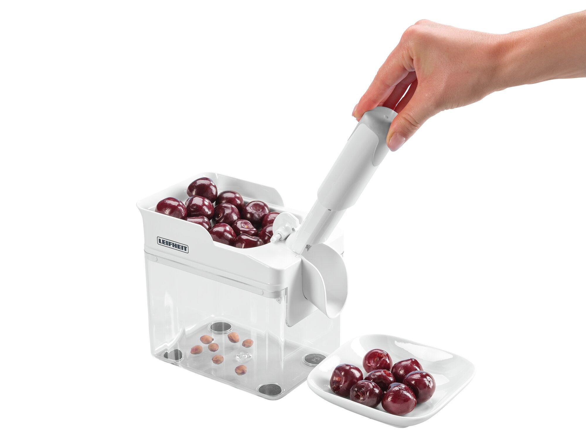 Kirschentkerner Cherrymat im Einsatz: Eine Hand bedient den weißen Kirschentkerner, der Kirschen entkernt und die Kerne in einem transparenten Behälter sammelt. Daneben ein Teller mit entkernten Kirschen.