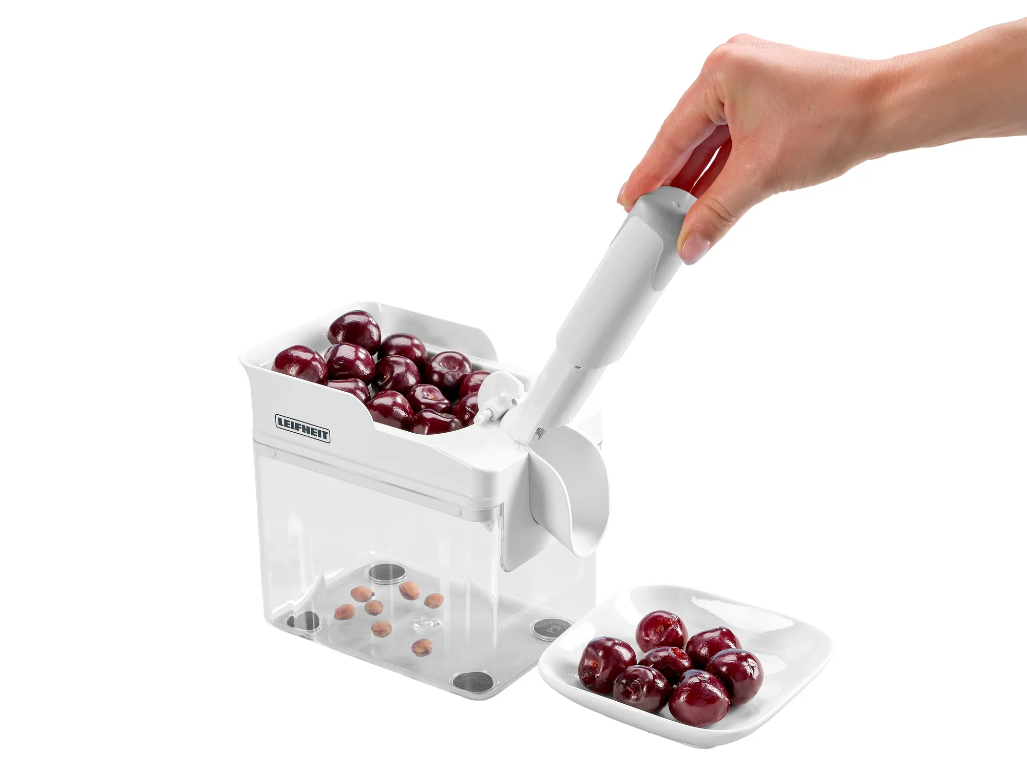Kirschentkerner Cherrymat im Einsatz: Eine Hand bedient den weißen Kirschentkerner, der Kirschen entkernt und die Kerne in einem transparenten Behälter sammelt. Daneben ein Teller mit entkernten Kirschen.