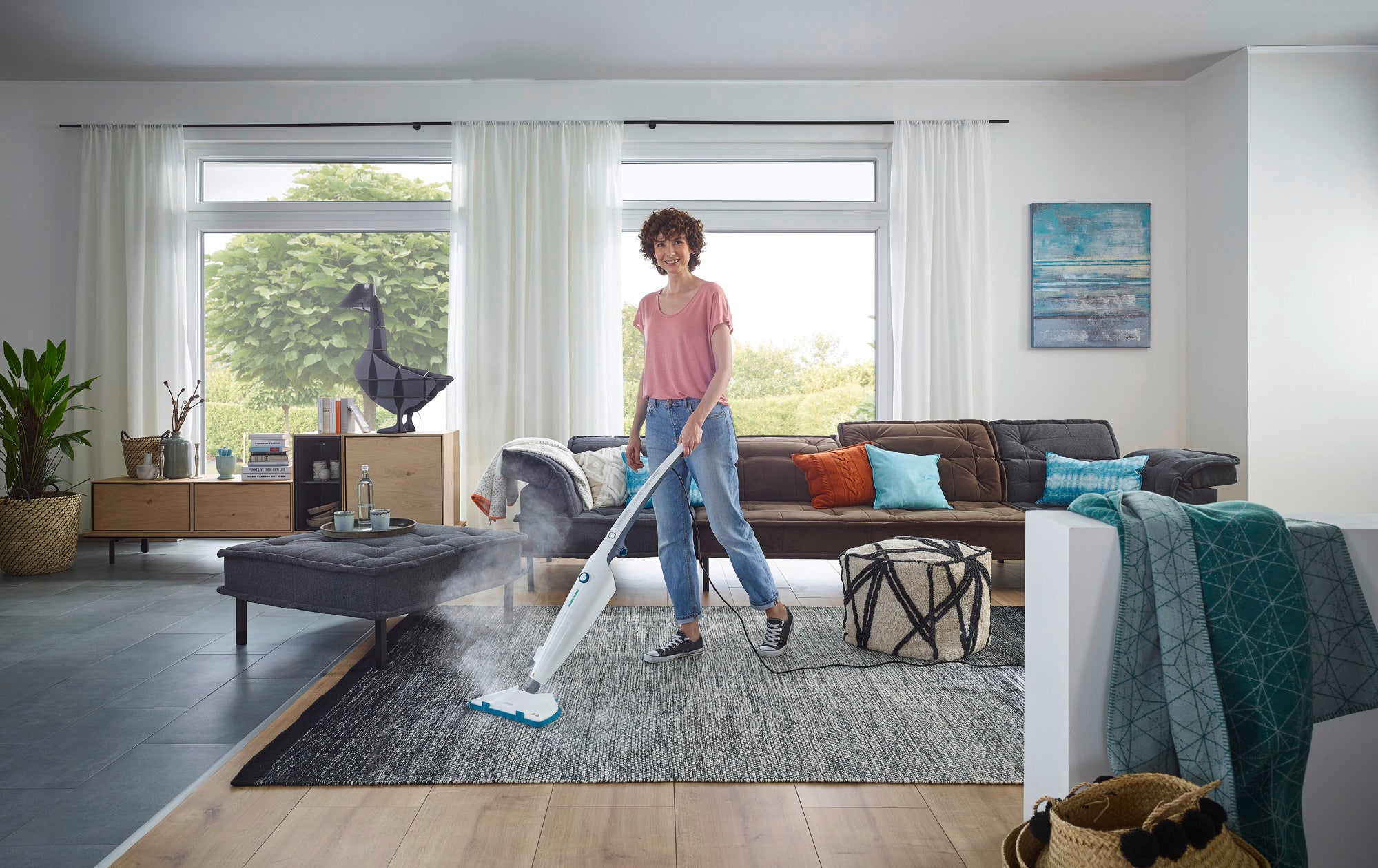 Une personne nettoie un tapis dans un salon moderne avec le nettoyeur vapeur CleanTenso Power. La pièce est lumineuse et élégamment meublée, avec de grandes fenêtres et des plantes.