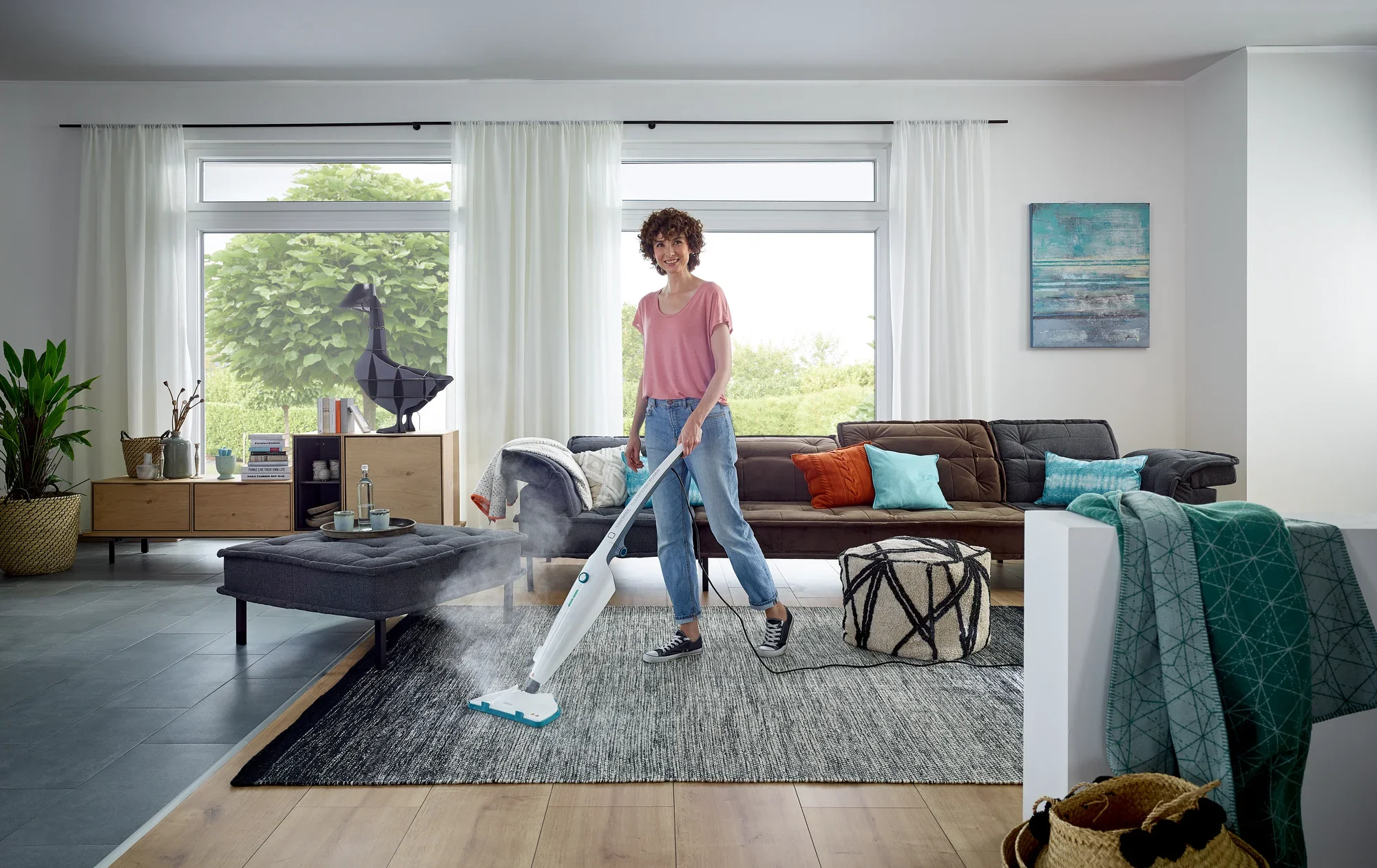 Une personne nettoie un tapis dans un salon moderne avec le nettoyeur vapeur CleanTenso Power. La pièce est lumineuse et élégamment meublée, avec de grandes fenêtres et des plantes.