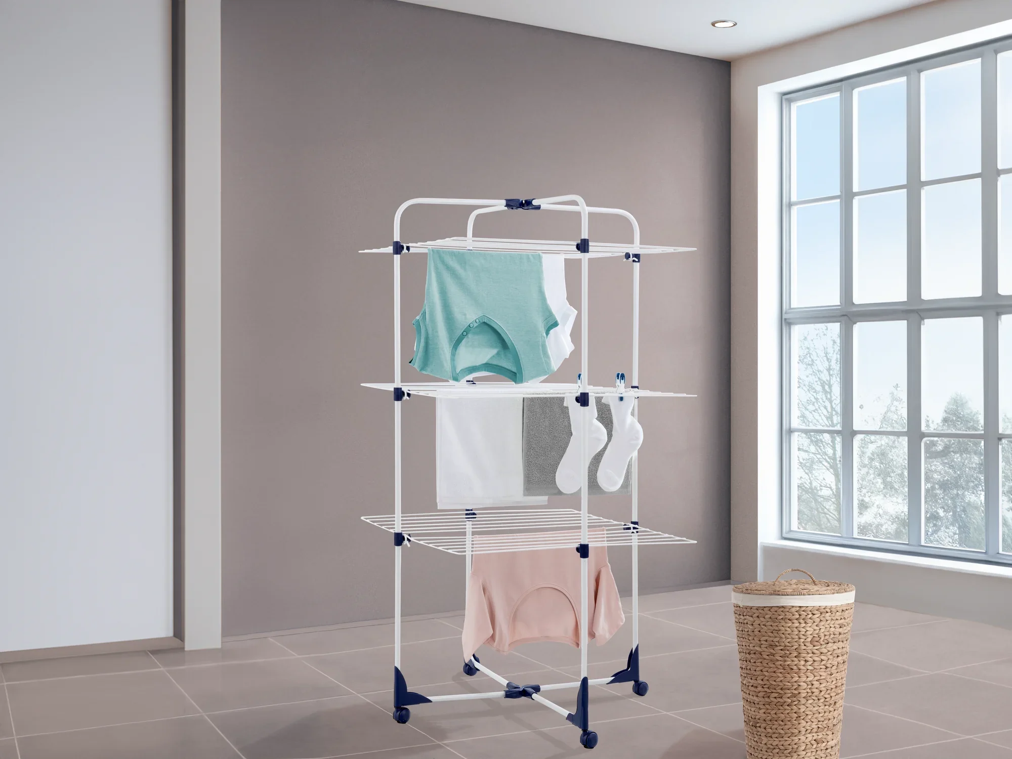 Een droogtoren met verschillende niveaus voor het drogen van kleding in een moderne kamer met grote ramen. Kleurrijke kledingstukken hangen aan de droger.