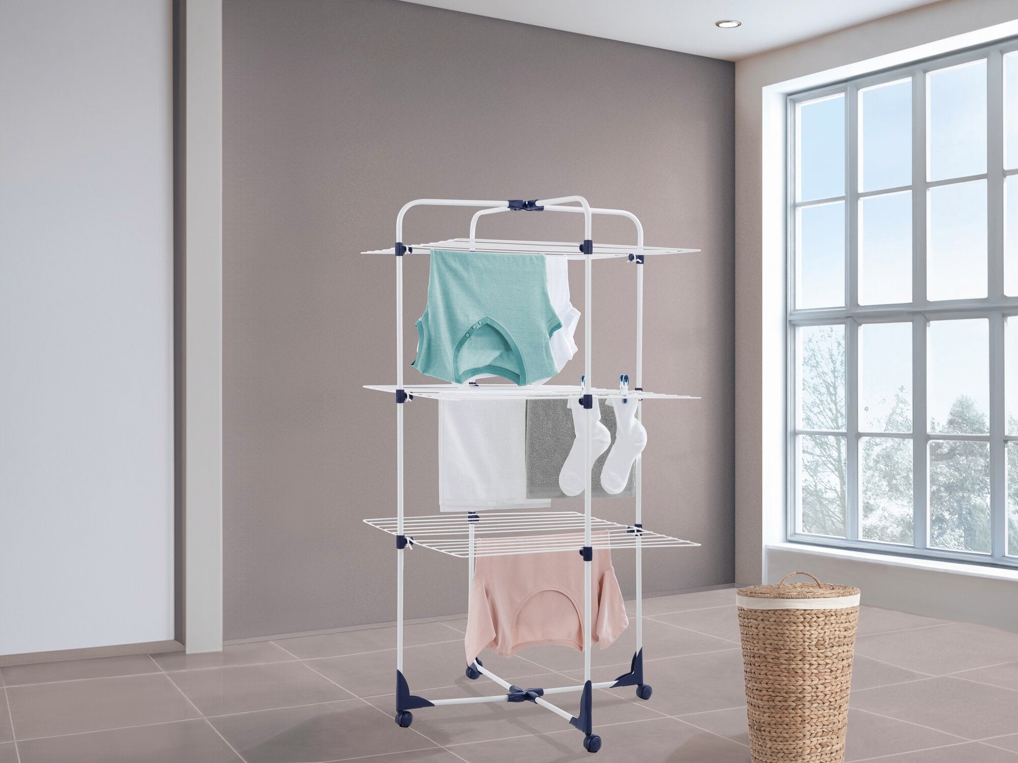 Een droogtoren met verschillende niveaus voor het drogen van kleding in een moderne kamer met grote ramen. Kleurrijke kledingstukken hangen aan de droger.