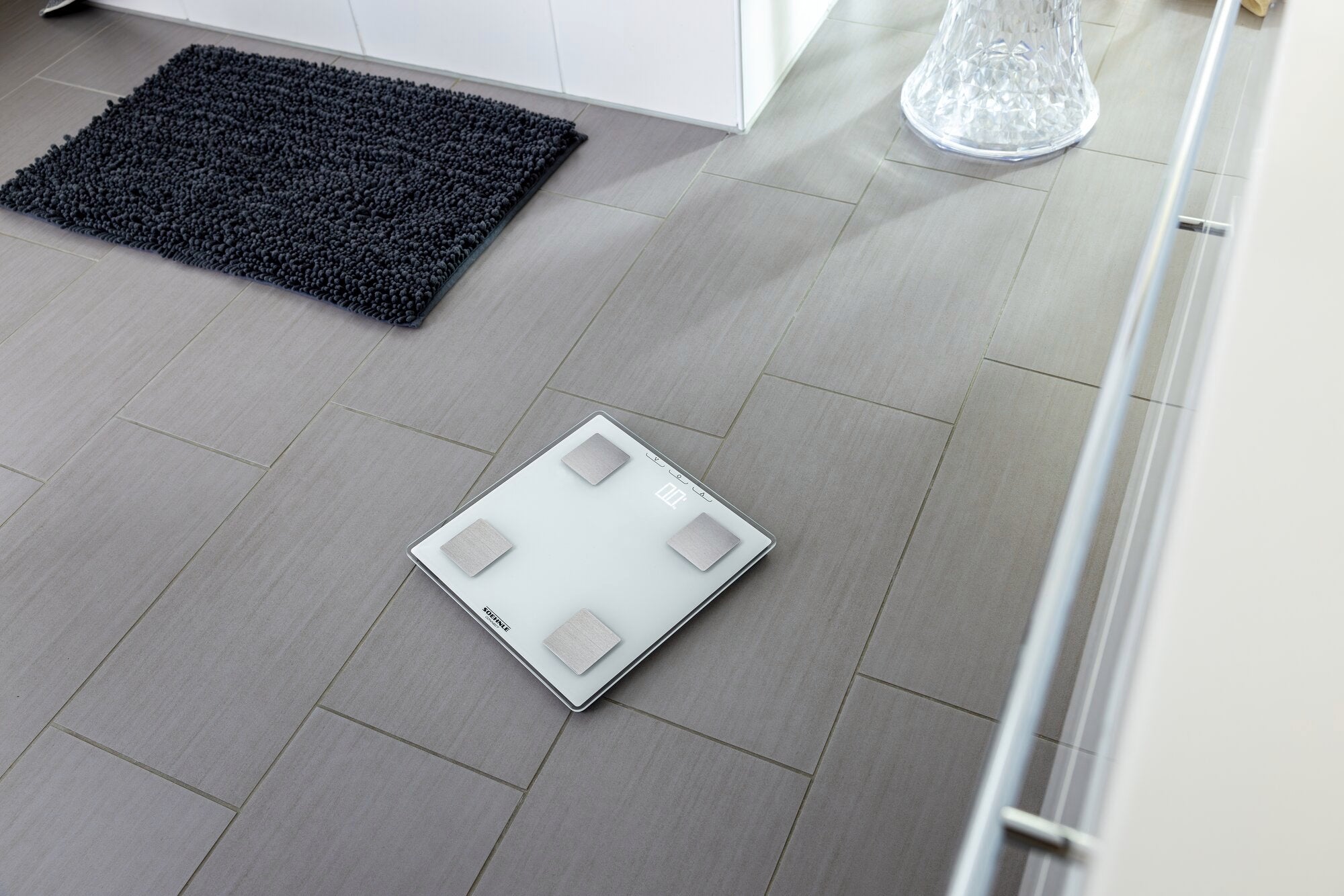 Eine digitale Personenwaage, Shape Sense Connect Lite, liegt auf einem modernen Badezimmerboden aus grauen Fliesen. Die Waage hat ein schlankes Design mit vier Sensoren.