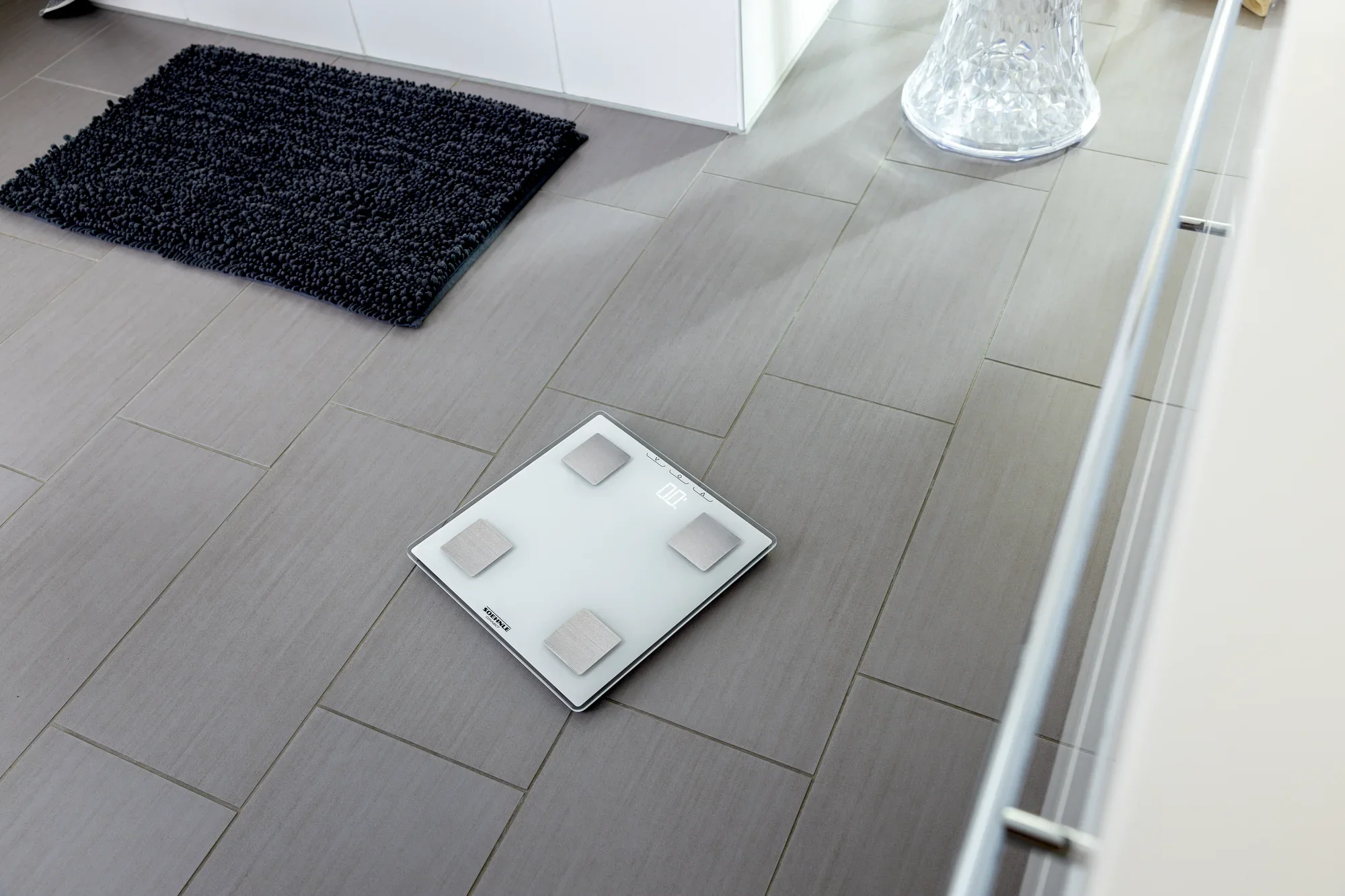 Cyfrowa waga łazienkowa Shape Sense Connect Lite leży na nowoczesnej podłodze łazienki wykonanej z szarych płytek. Waga ma elegancką konstrukcję z czterema czujnikami.