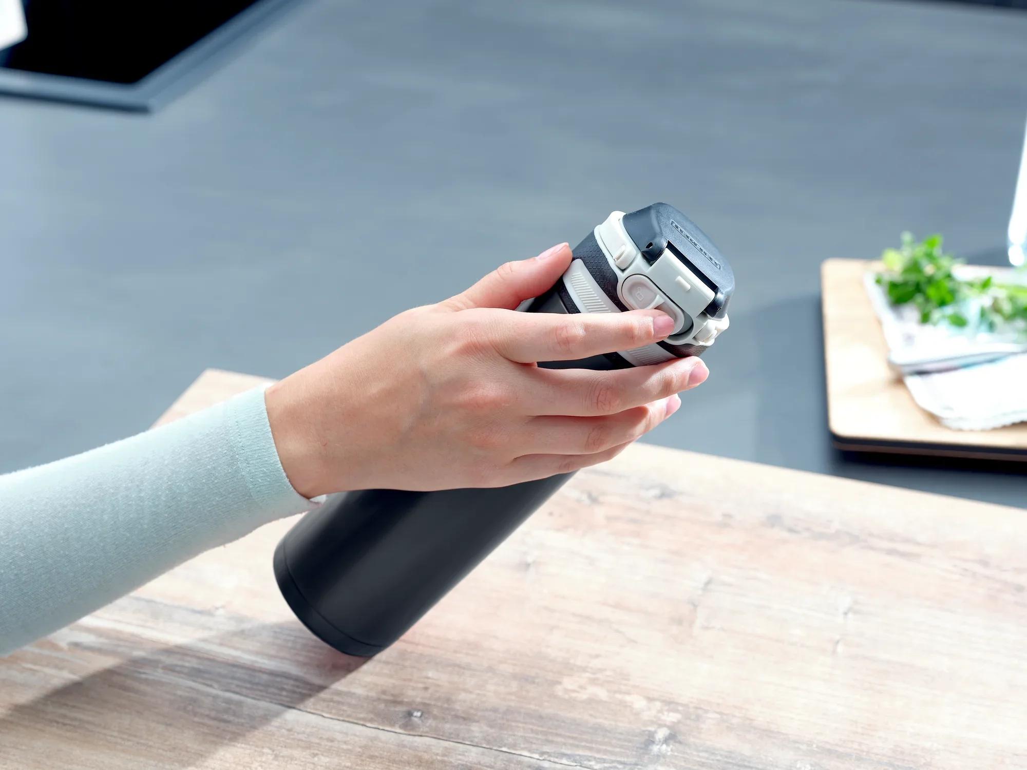 Izolowany kubek Flip 600 ml w kolorze czarnym jest trzymany jedną ręką na drewnianym stole. Kubek ma praktyczną pokrywkę typu flip-top, idealną w podróży.