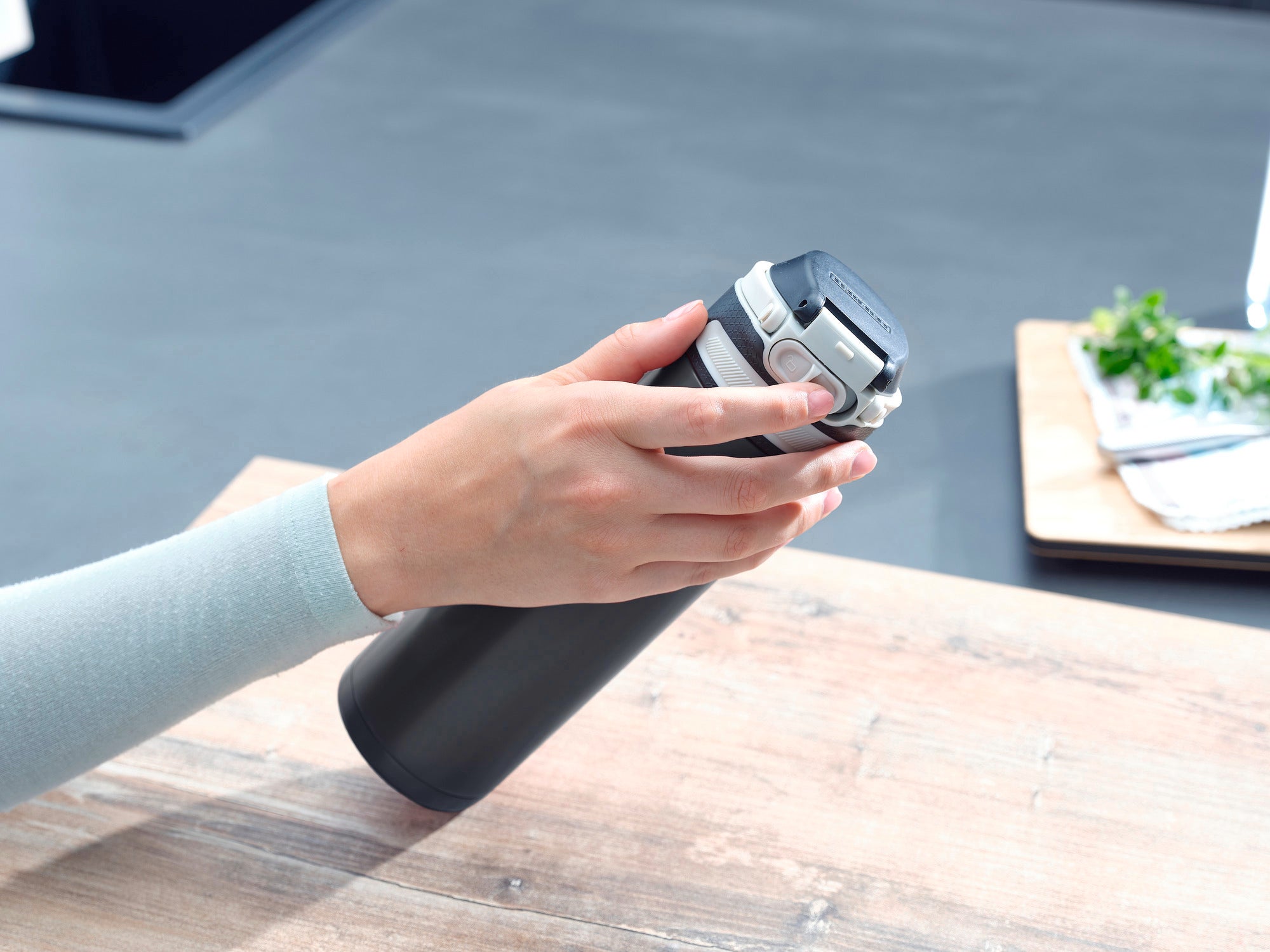 Izolowany kubek Flip 600 ml w kolorze czarnym jest trzymany jedną ręką na drewnianym stole. Kubek ma praktyczną pokrywkę typu flip-top, idealną w podróży.