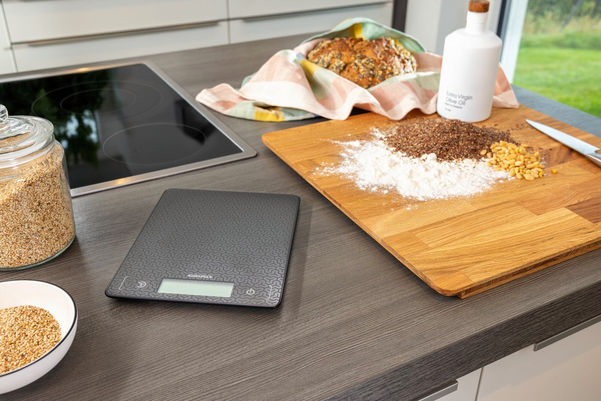 Digitale Küchenwaage Comfort Mosaic auf einer Küchenarbeitsplatte neben Zutaten wie Mehl, Körnern und Brot. Perfekt für präzises Abwiegen beim Backen und Kochen.