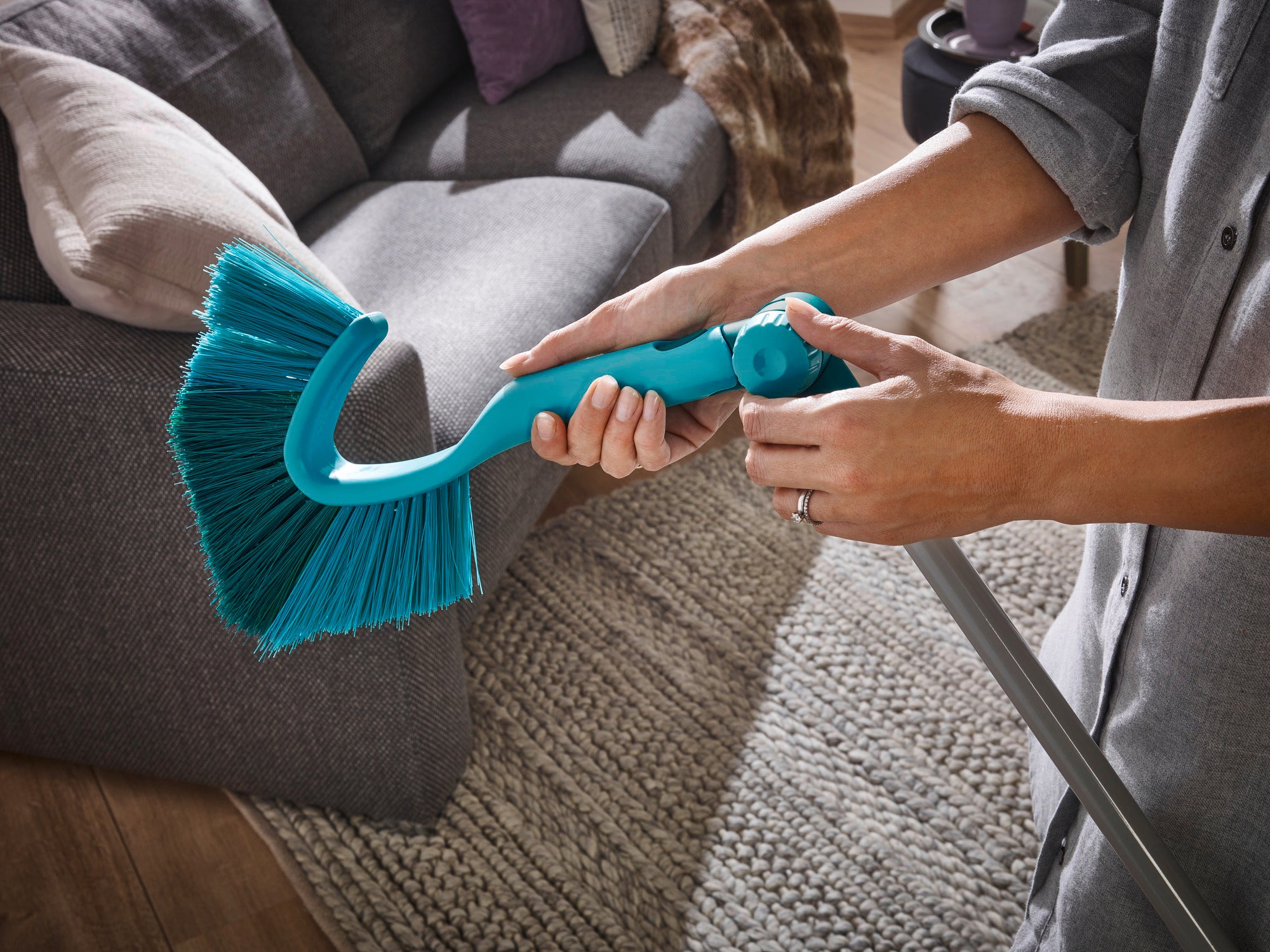 A person is holding a dustpan and brush with a telescopic handle in a cosy living room. The brush has blue bristles and is ideal for hard-to-reach places.