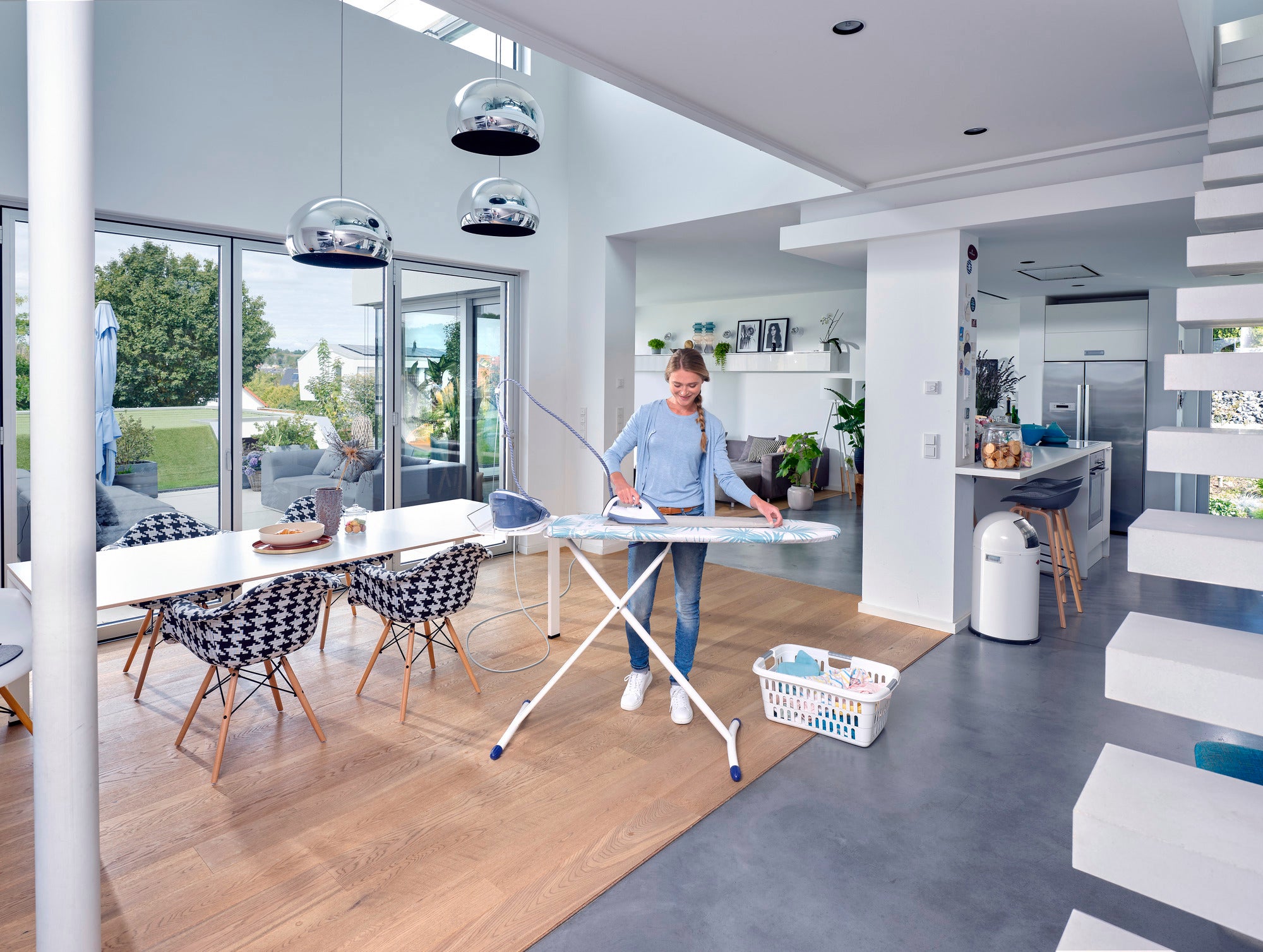 Air Board Express M Stevige strijkplank in een moderne, lichte kamer. Eén persoon strijkt kleding terwijl het daglicht door de grote ramen schijnt. Een wasmand staat ernaast.