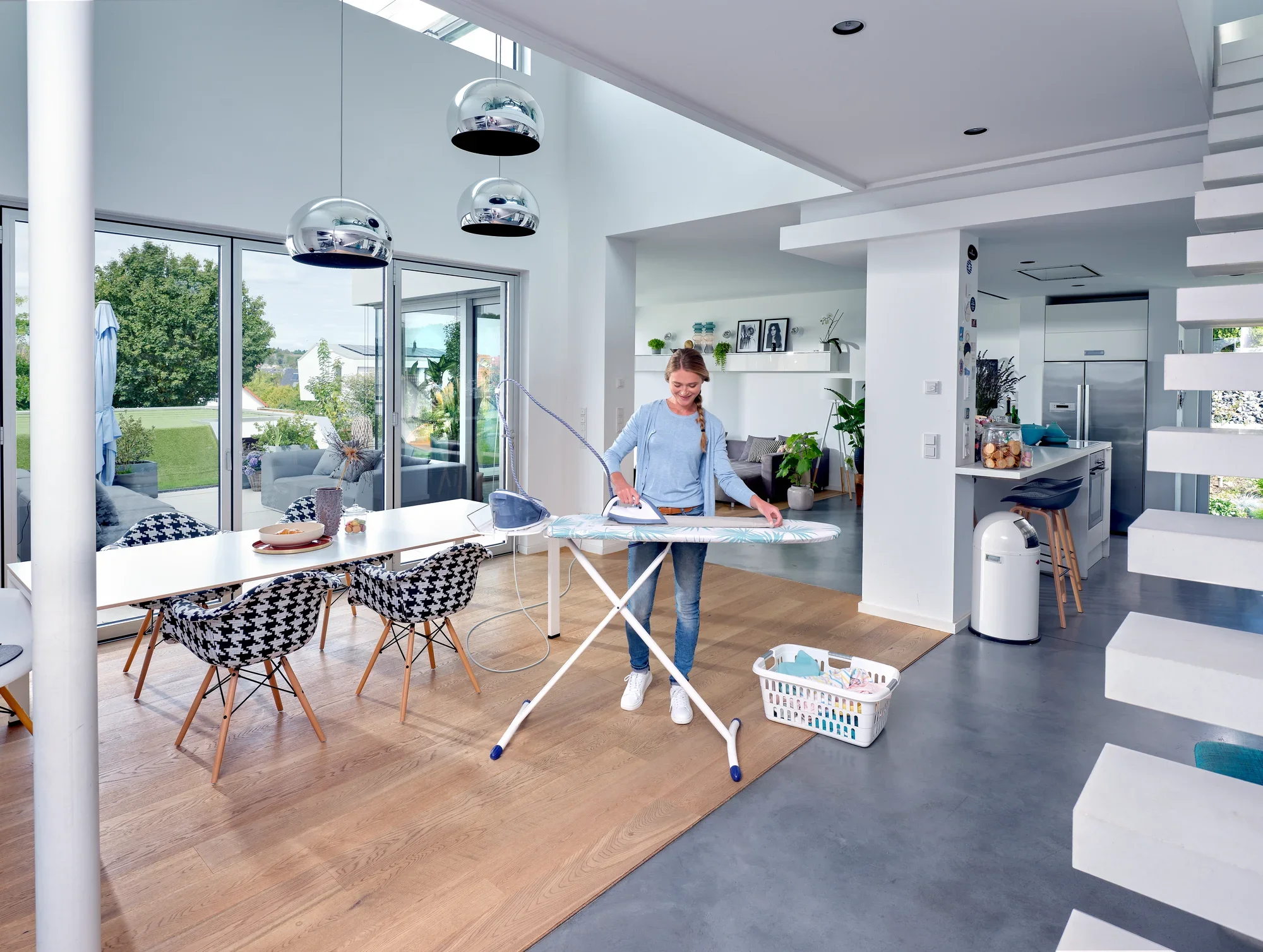 Table à repasser Air Board Express M Solid dans une pièce moderne et lumineuse. Une personne repasse des vêtements tandis que la lumière du jour pénètre par de grandes fenêtres. Un panier à linge se trouve à côté.