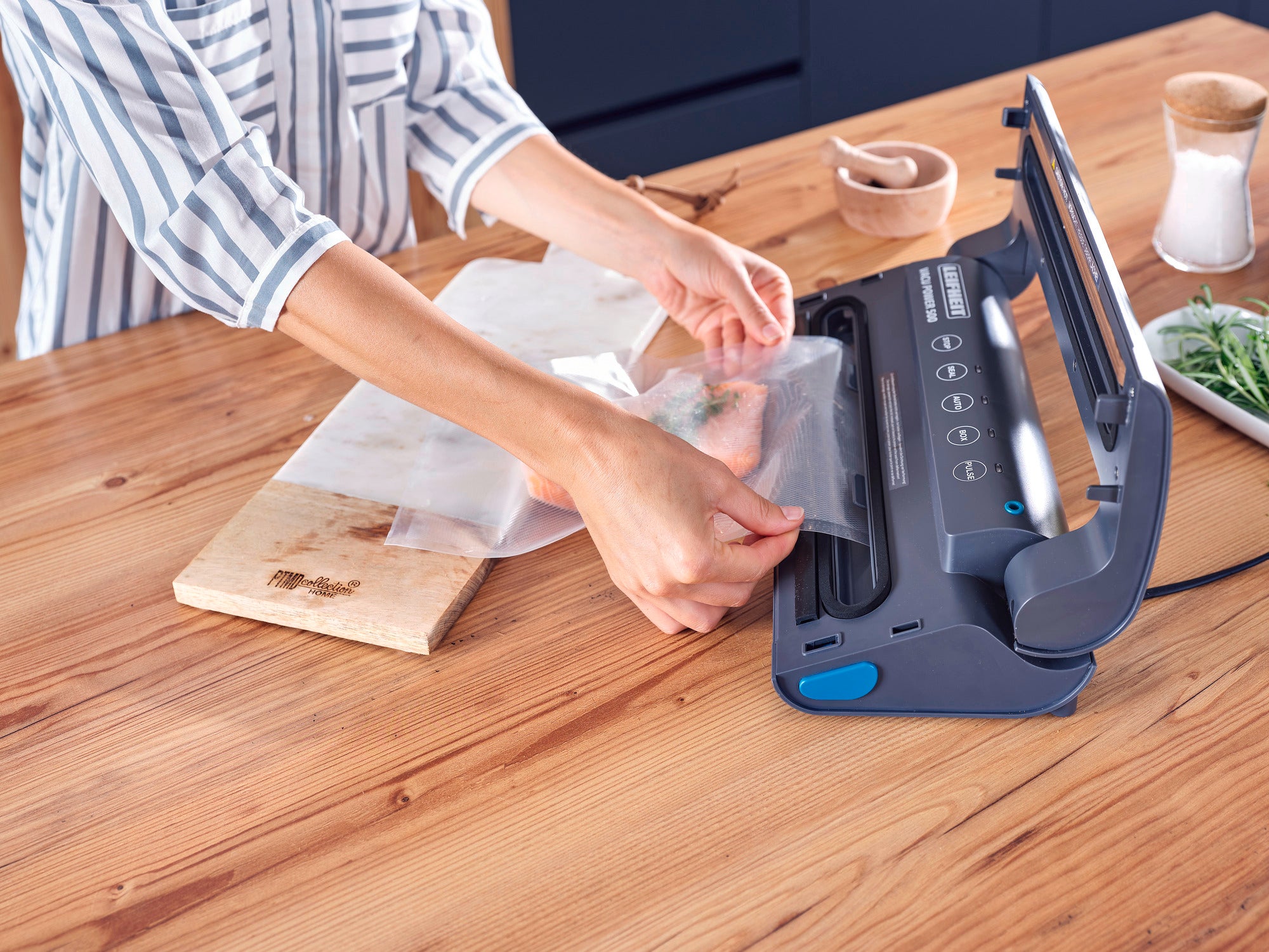 Eine Person verwendet das Vakuumiergerät Vacu Power 500, um Lebensmittel in einem Beutel zu versiegeln. Das Gerät liegt auf einem Holztisch in einer modernen Küche.