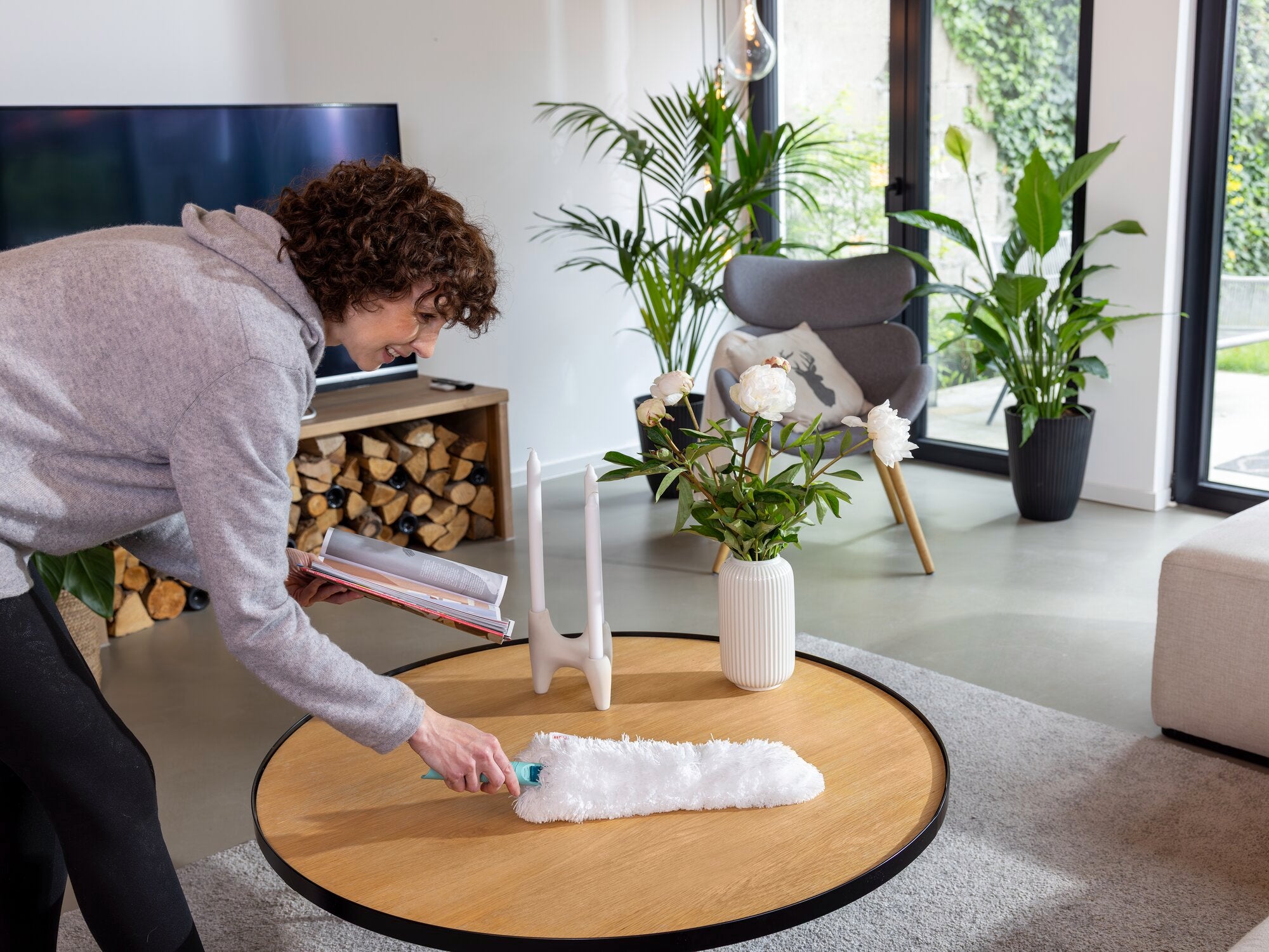 Eine Person verwendet den SUPERDUSTER XL Staubwedel, um einen Tisch in einem modernen Wohnzimmer zu reinigen. Im Hintergrund sind Pflanzen und ein Kamin zu sehen.