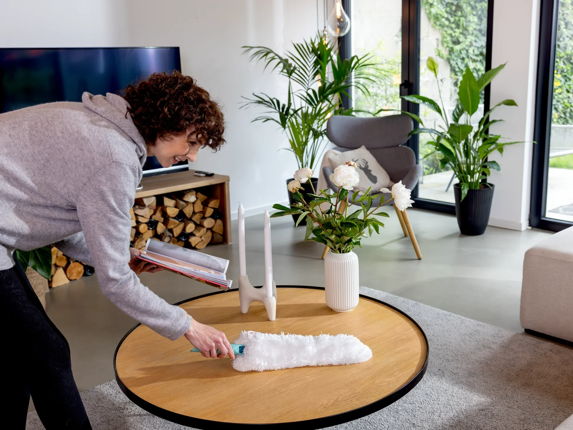 Una persona utiliza el plumero SUPERDUSTER XL para limpiar una mesa en un salón moderno. Al fondo se ven plantas y una chimenea.