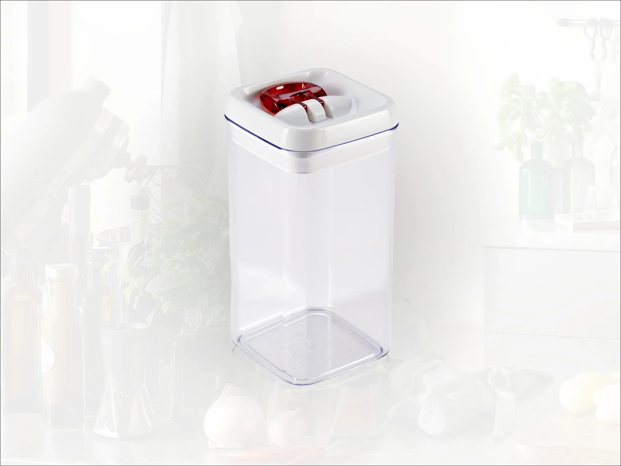Un recipiente transparente y rectangular con tapa roja. Ideal para guardar alimentos en la cocina. Al fondo se ven utensilios de cocina.
