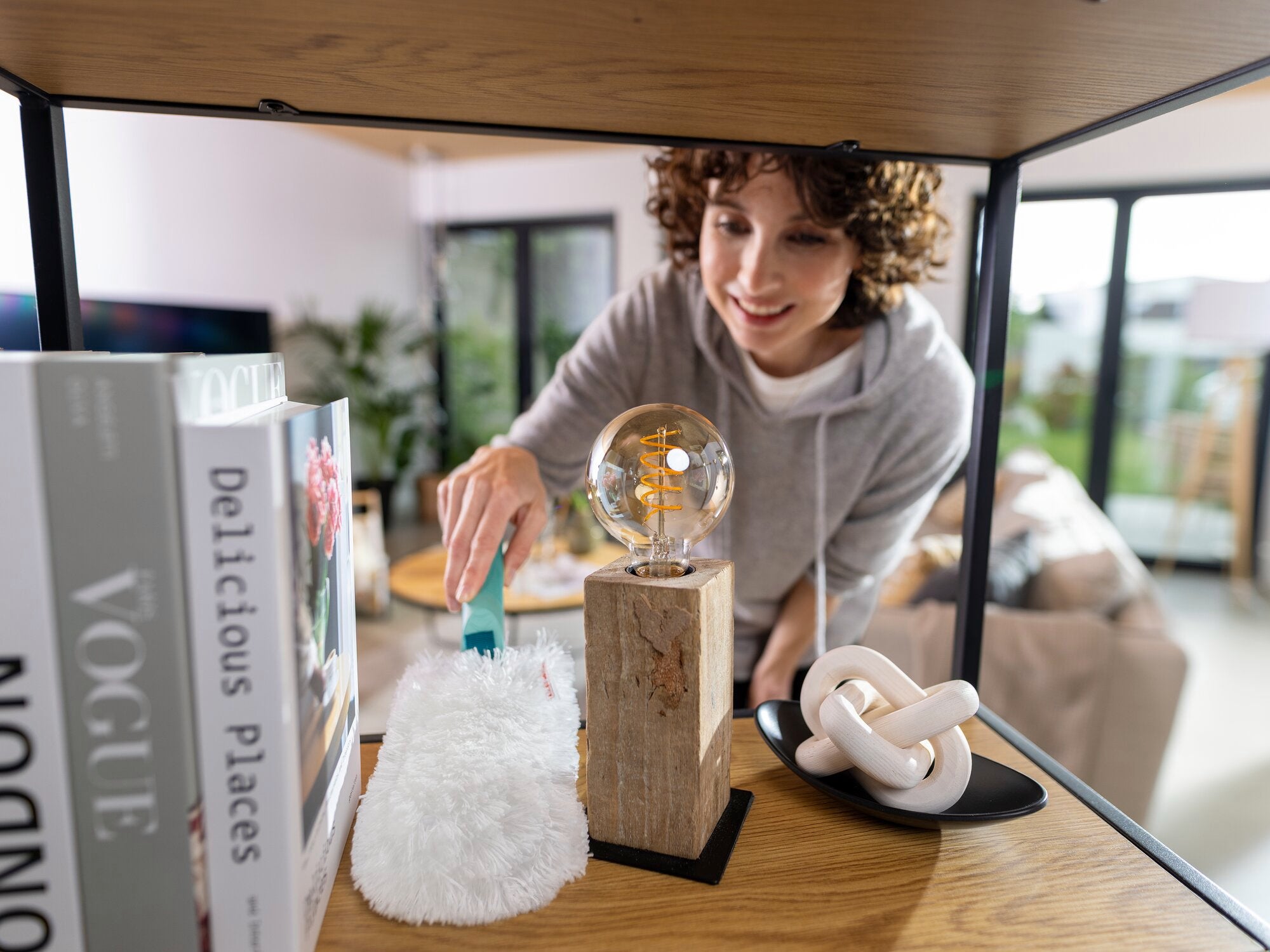 Une personne utilise le plumeau SUPERDUSTER pour nettoyer une étagère. Le plumeau est moelleux et blanc, idéal pour enlever la poussière sur les surfaces délicates.