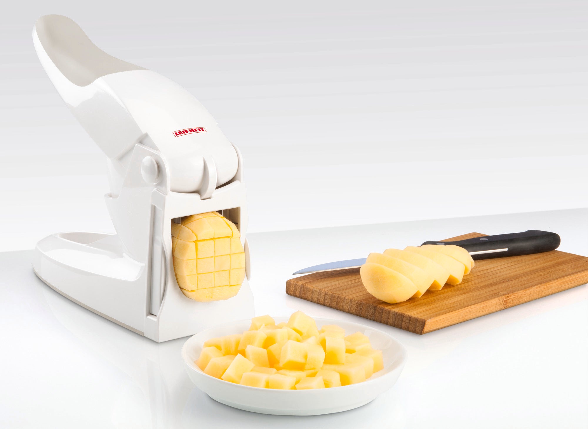 Pommes-Frites-Schneider in Aktion: Ein weißer Schneider schneidet eine Kartoffel in gleichmäßige Stäbchen. Im Hintergrund liegt eine geschnittene Kartoffel auf einem Holzbrett.