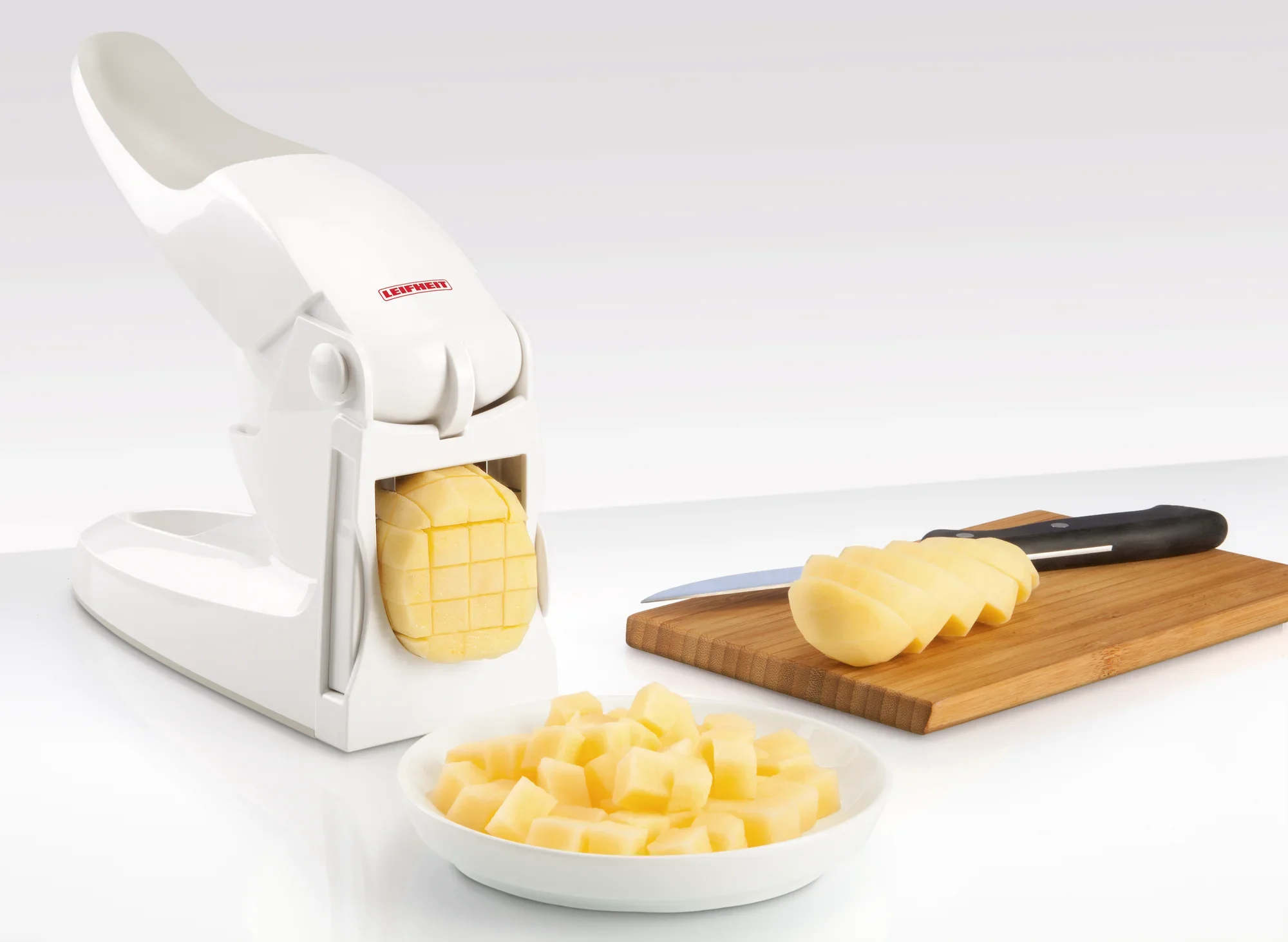 Fritessnijder in actie: Een witte snijder snijdt een aardappel in gelijkmatige staafjes. Op de achtergrond ligt een gesneden aardappel op een houten plank.