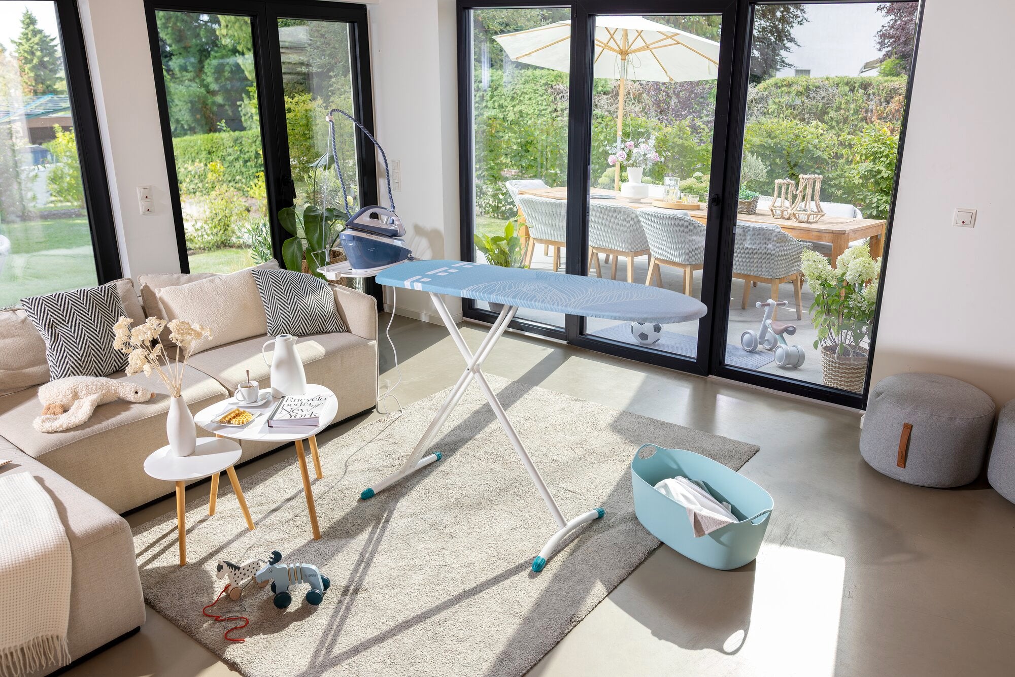 Bügelbrett Leifheit Air Board Express M Solid in einem modernen Wohnzimmer. Es steht auf einem Teppich, umgeben von Möbeln und großen Fenstern mit Blick auf einen Garten.