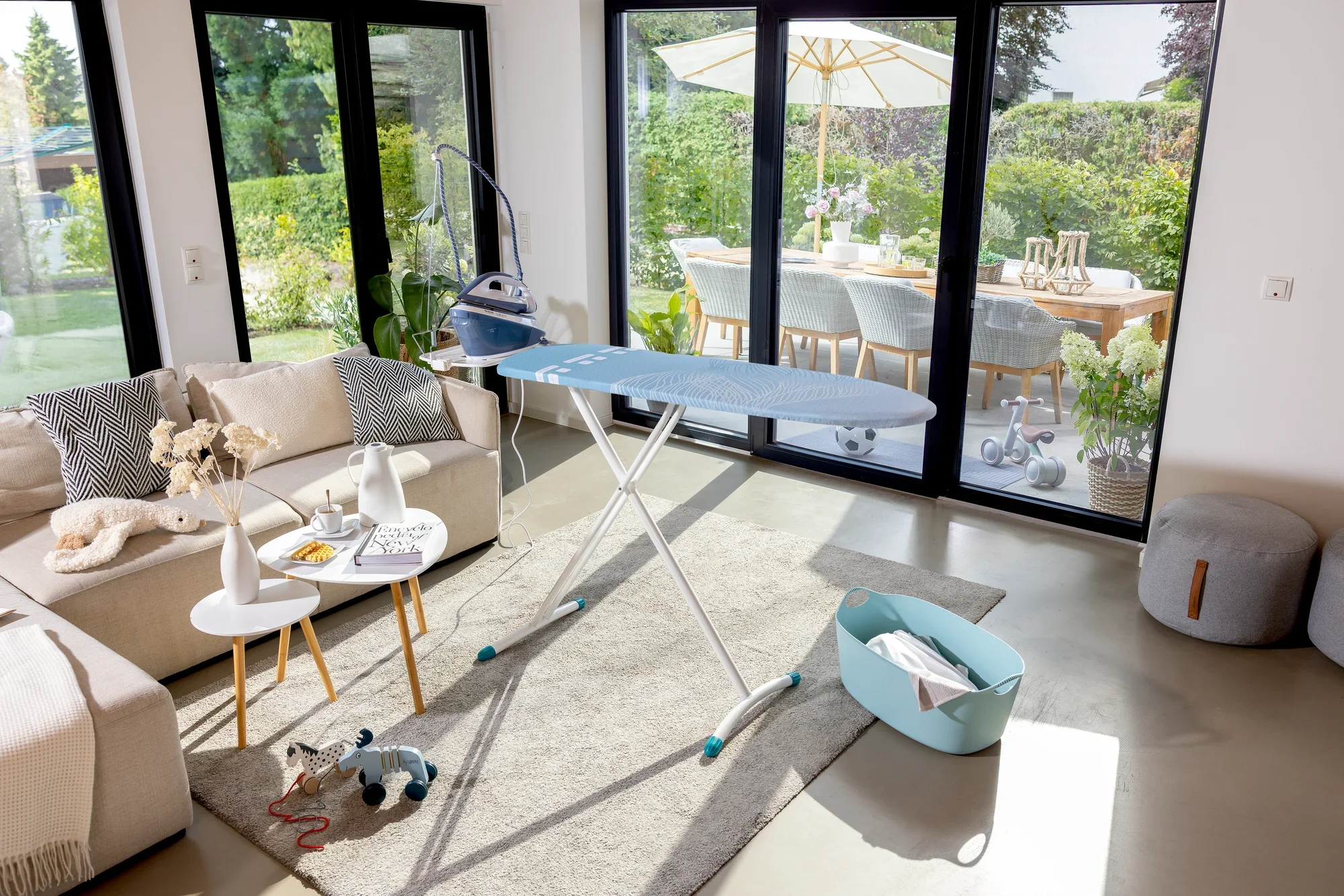 Bügelbrett Leifheit Air Board Express M Solid in einem modernen Wohnzimmer. Es steht auf einem Teppich, umgeben von Möbeln und großen Fenstern mit Blick auf einen Garten.