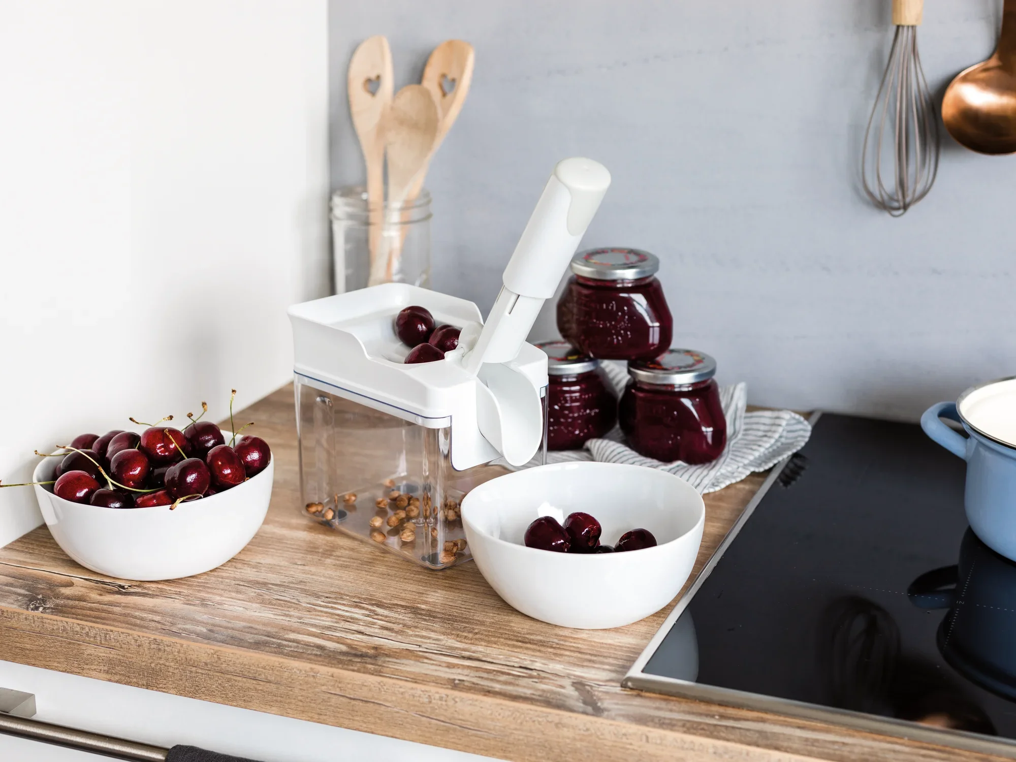 Kirschentkerner Cherrymat auf einer Küchenarbeitsplatte, umgeben von Schalen mit frischen Kirschen und Gläsern mit Kirschmarmelade. Ideal zum schnellen Entkernen von Kirschen.