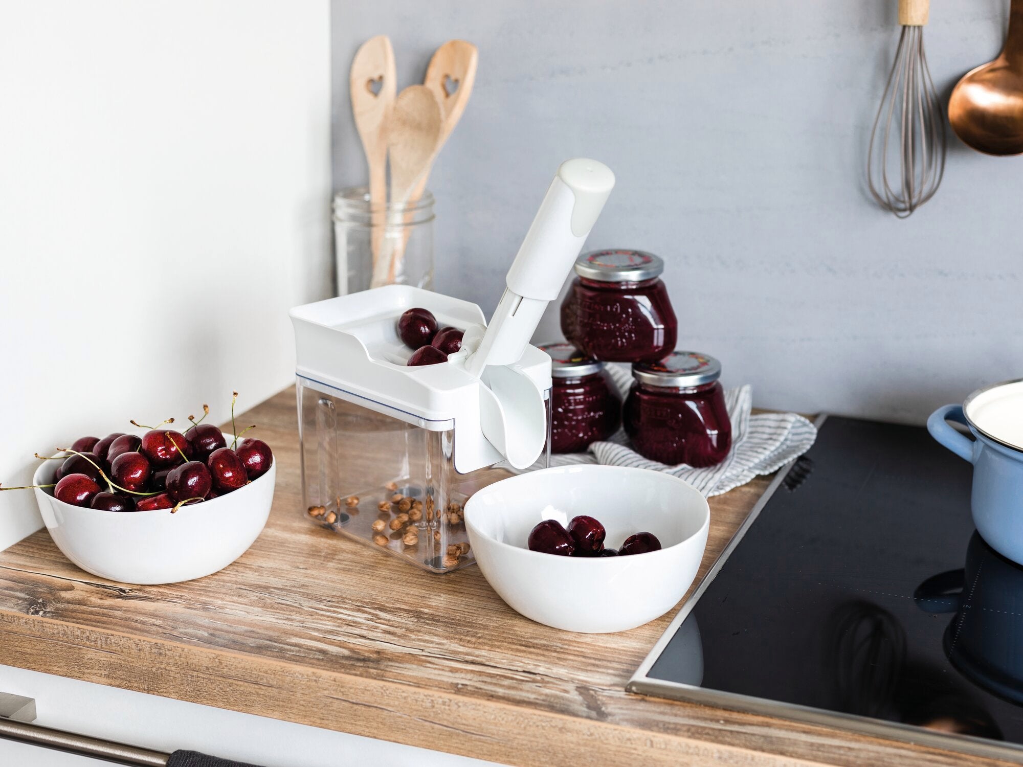 Drylownica do wiśni Cherrymat na blacie kuchennym, otoczona miskami świeżych wiśni i słoikami dżemu wiśniowego. Idealny do szybkiego drylowania wiśni.
