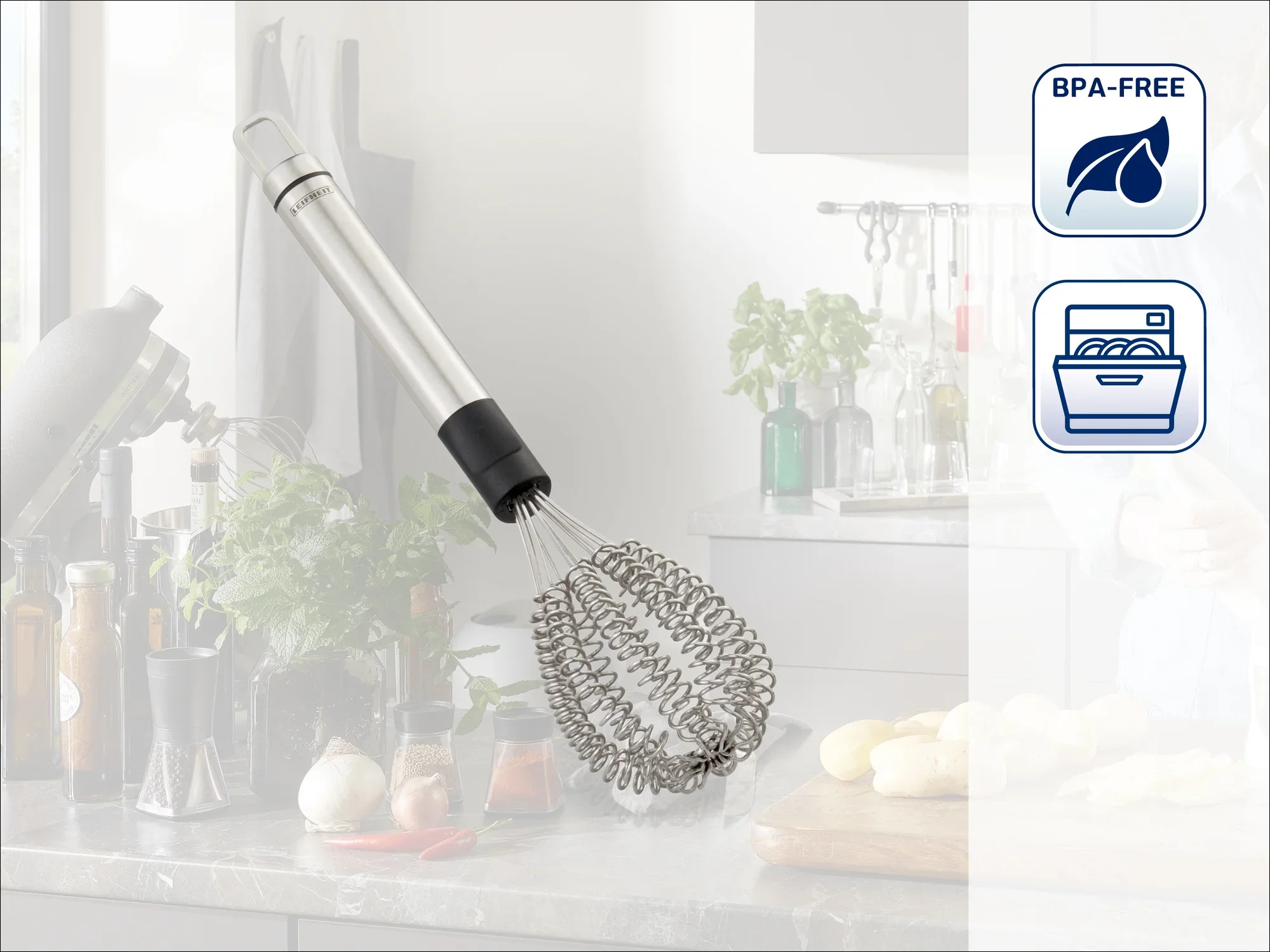 Un fouet en acier inoxydable avec un manche noir flotte devant une cuisine moderne. À l'arrière-plan, on aperçoit des ingrédients frais et des ustensiles de cuisine. L'image montre les symboles « sans BPA » et « lavable au lave-vaisselle ».