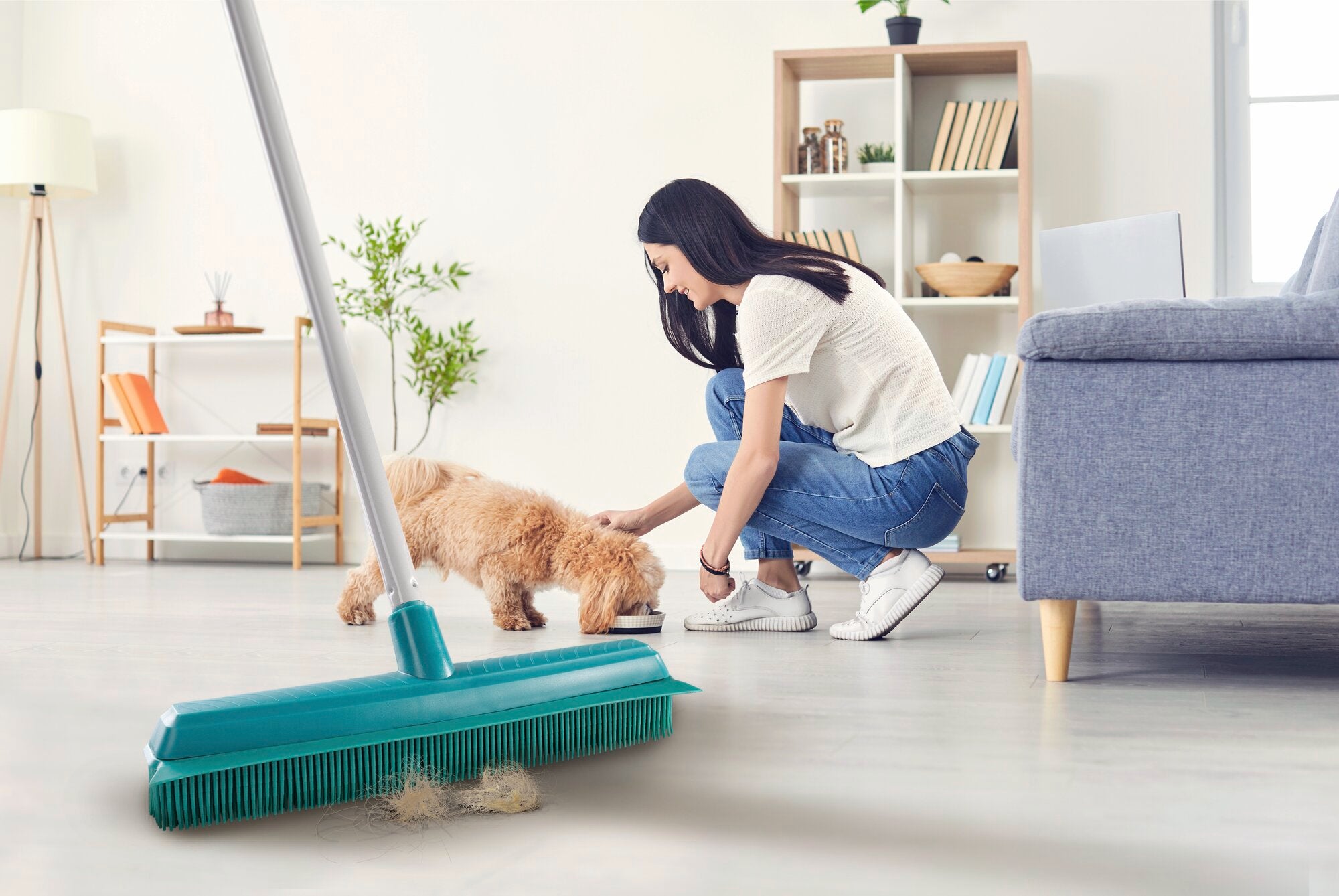 Frau reinigt den Boden mit dem Leifheit Suprabesen, während ein Hund in einem modernen Wohnzimmer frisst. Der Besen entfernt effektiv Schmutz und Tierhaare.